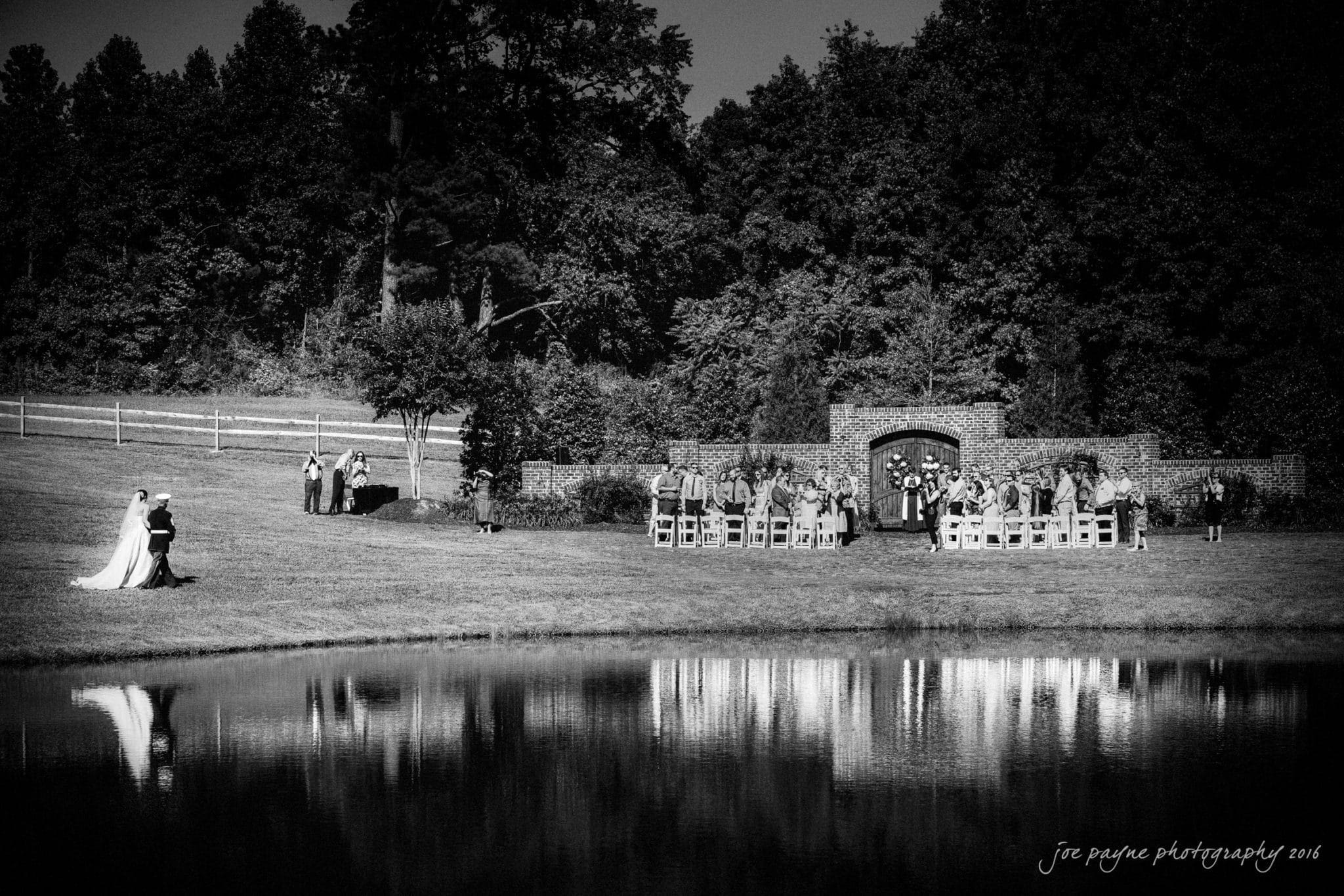 the oaks at salem wedding photography - nicole & brendan A black and white photo of an outdoor wedding ceremony by a lake. The bridal party is near the altar, and a couple stands apart, embracing. Trees and a brick arch form the backdrop, reflecting in the water.