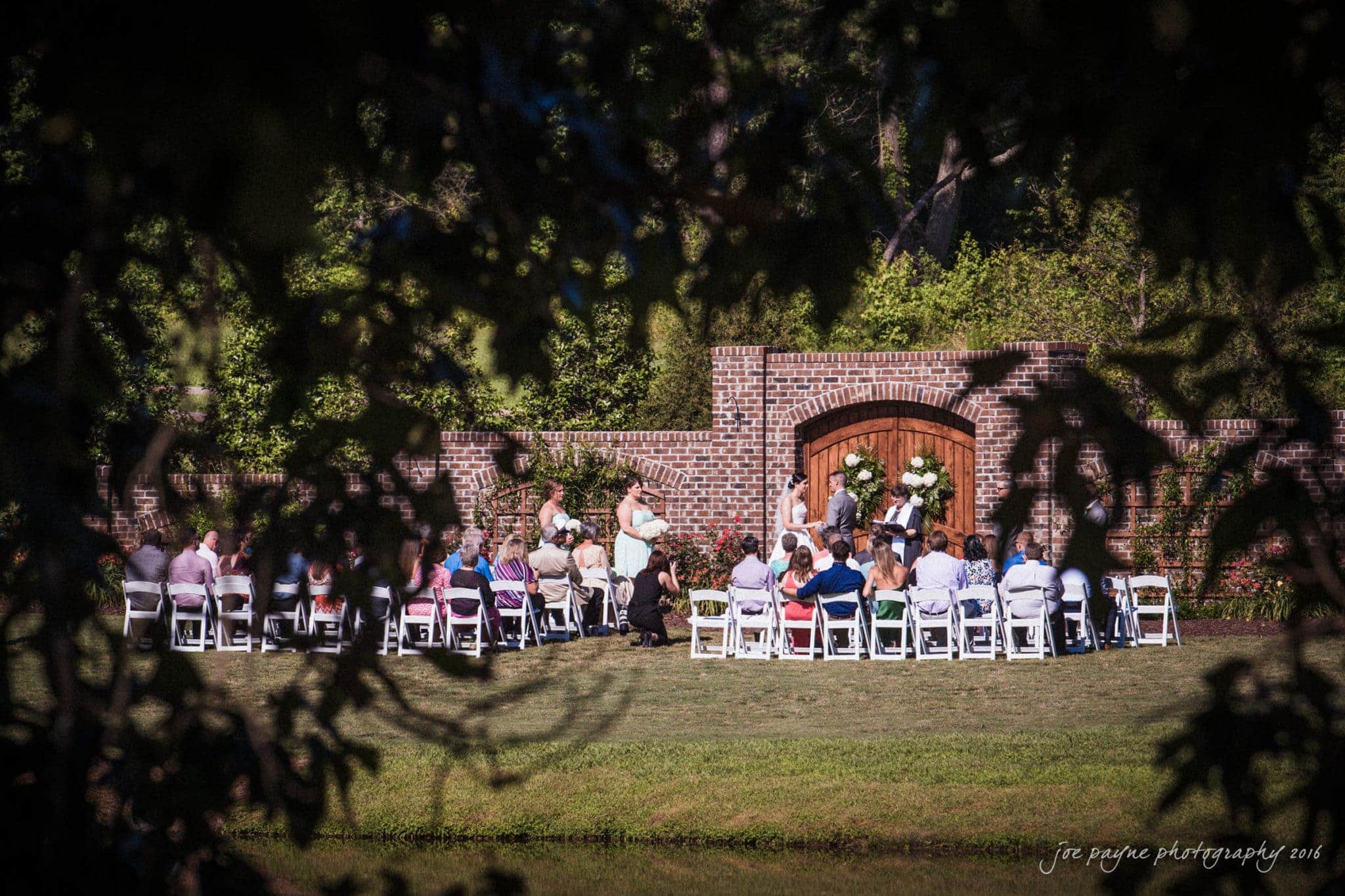the oaks at salem wedding photography - nicole & brendan A wedding ceremony takes place outdoors. Guests are seated on white chairs facing a brick arch adorned with flowers. Trees and greenery frame the scene, creating a natural, intimate setting.
