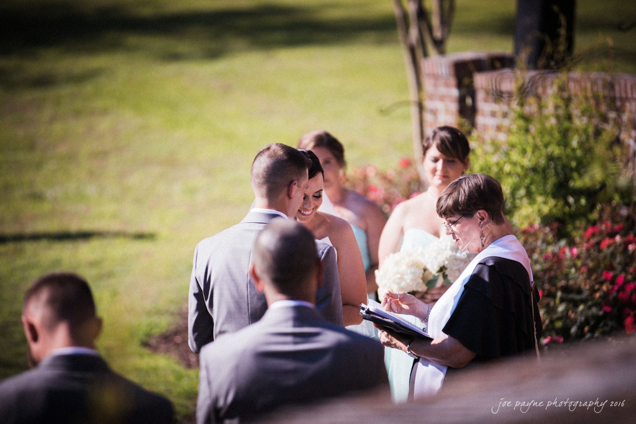 the oaks at salem wedding photography - nicole & brendan A wedding ceremony outdoors with the bride and groom facing a minister. Bridesmaids hold bouquets in the background, and the setting is lush with greenery and flowers.