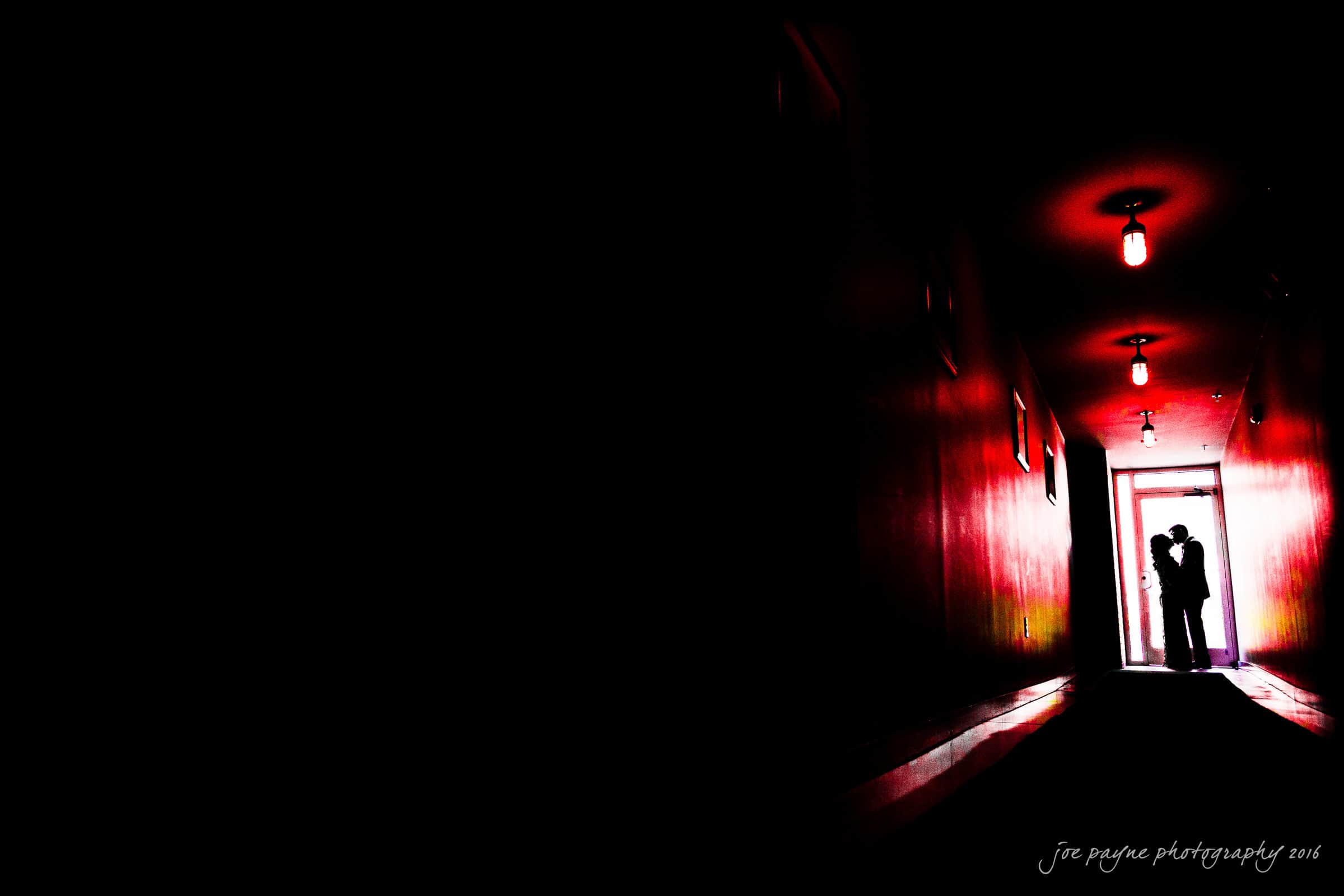 A dimly lit hallway with red walls leads to a silhouetted couple embracing in a doorway at the end. A single overhead light casts a warm glow, creating a dramatic and romantic atmosphere.