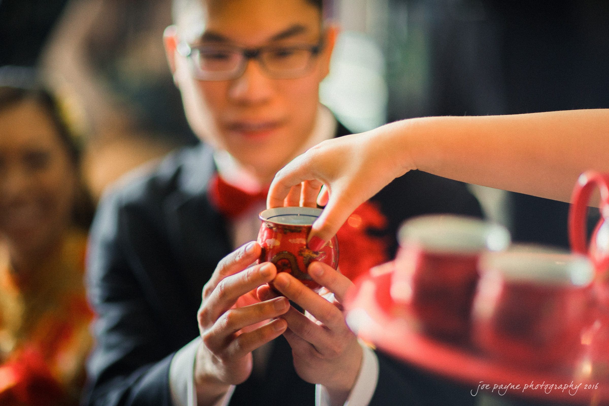 A person in formal attire holds a red teacup with intricate designs as another hand offers it to him. Nearby are more red teacups on a tray, suggesting a traditional tea ceremony setting.