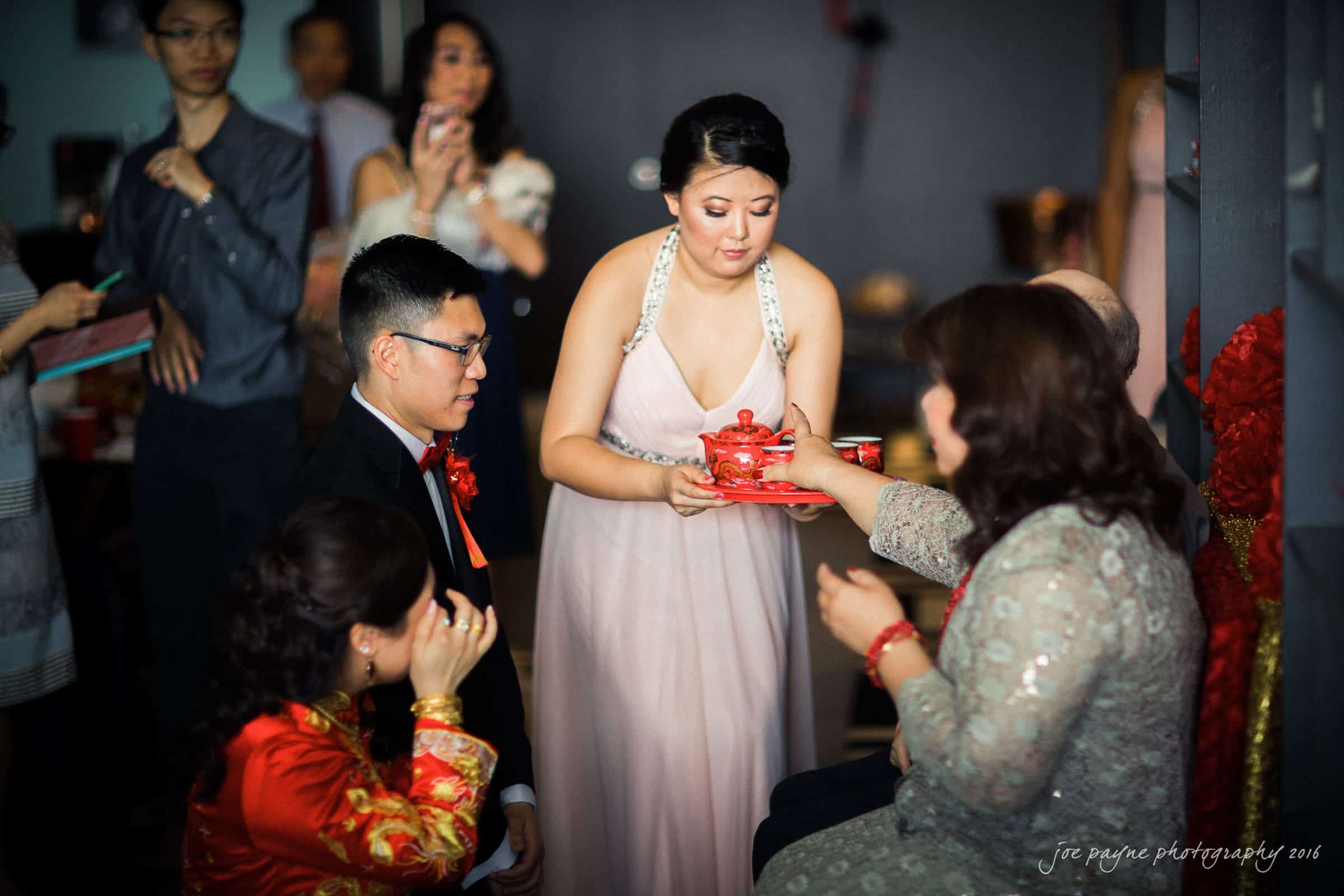 A woman in a light pink dress serves tea to seated guests, including a man in a black suit with a red tie, during a traditional Asian tea ceremony. Other people are gathered in the background, observing the ritual.