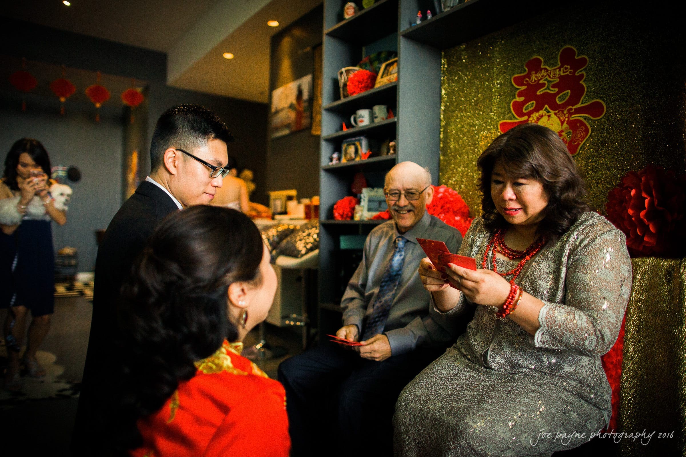 A couple in traditional attire kneels before an older man and woman seated, who are giving them red envelopes. The room is decorated with red and gold ornaments, symbolizing a cultural celebration. Others are visible in the background.