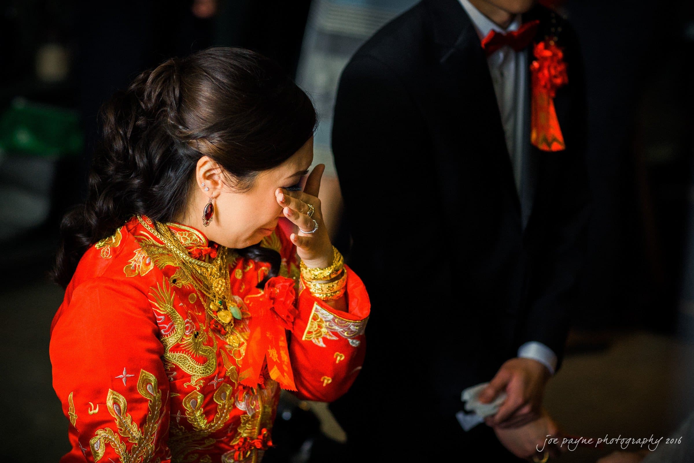 A woman in a red and gold traditional gown wipes a tear from her eye, holding a tissue. She wears gold jewelry and the background features a person in a black suit with a red bow tie. The scene is emotional and intimate.