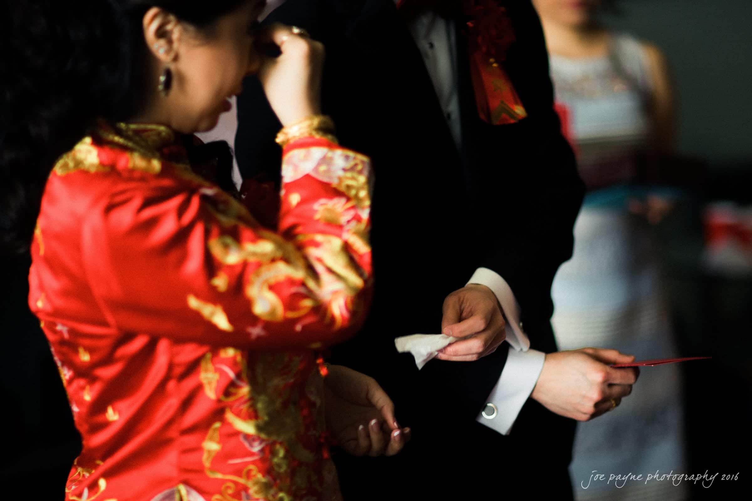 A woman in a traditional red dress wipes her eye while holding a tissue. A man beside her in a dark suit offers a handkerchief. A blurred person in the background watches.