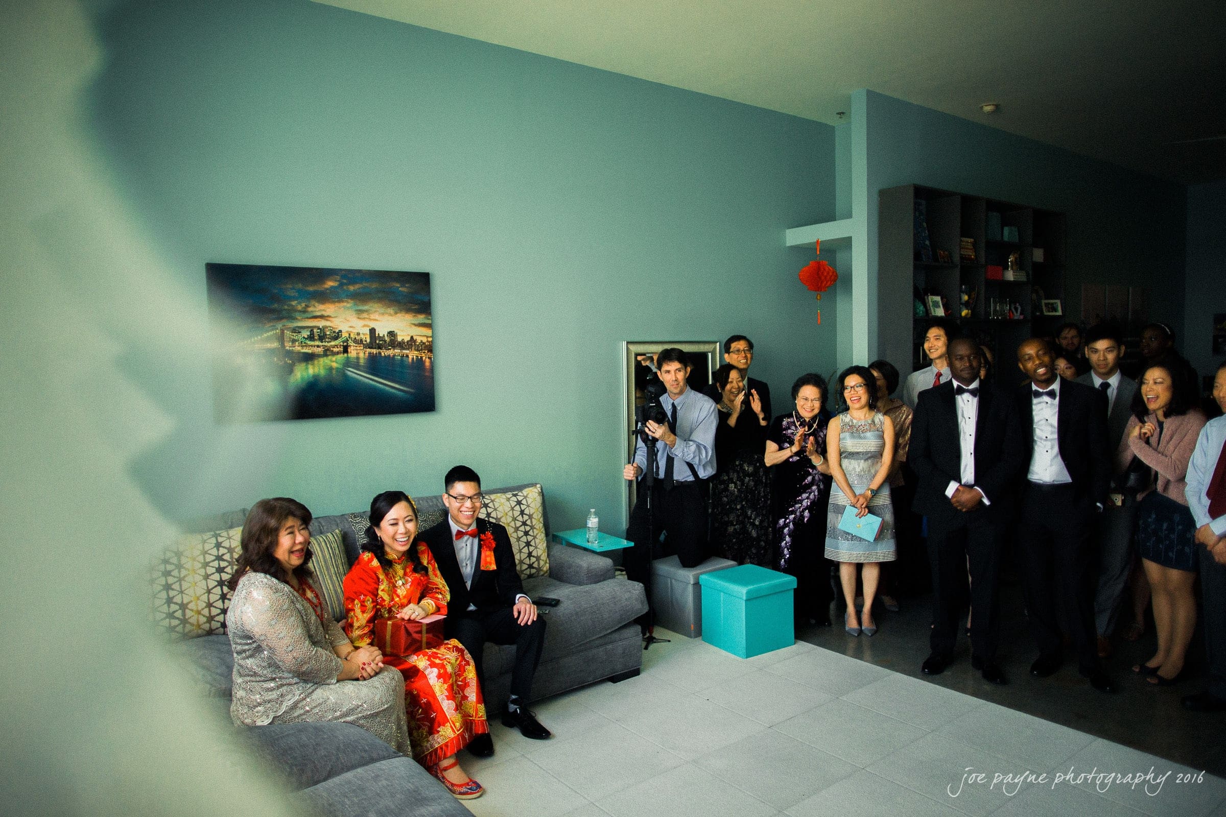 A group of people in formal attire gather in a living room. The couple on the left sits on a sofa wearing traditional clothing, surrounded by standing guests who appear joyful and attentive. The room is decorated with a cityscape painting and a red ornament.