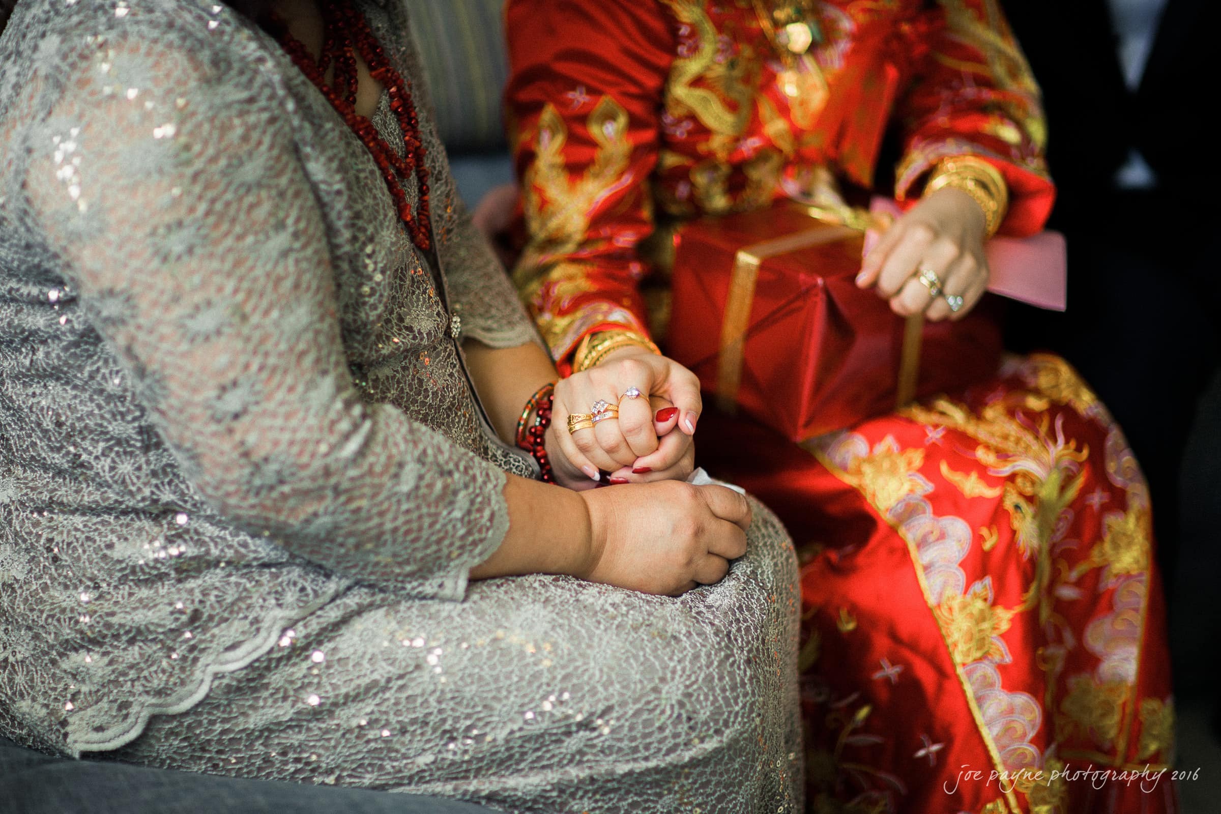 Two people dressed in ornate outfits are sitting close together. The person on the left wears a silver lace dress and holds hands with the person on the right, who wears a red and gold dress, and holds a wrapped gift.