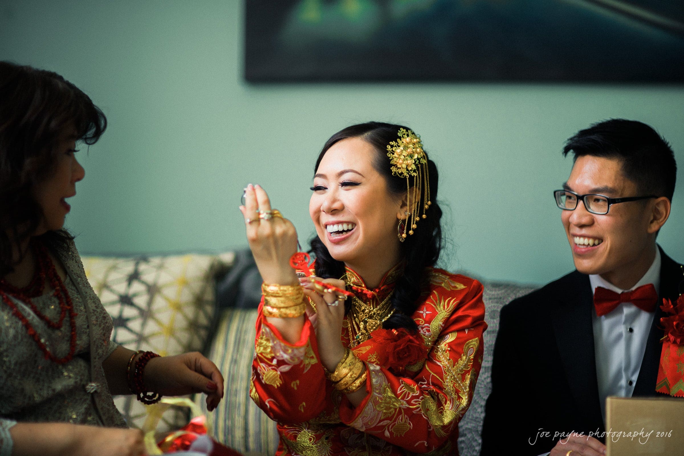 A woman in a red and gold traditional dress smiles and shows her rings, sitting between a woman and a man in formal attire. The group is indoors, on a couch, with pillows in the background.