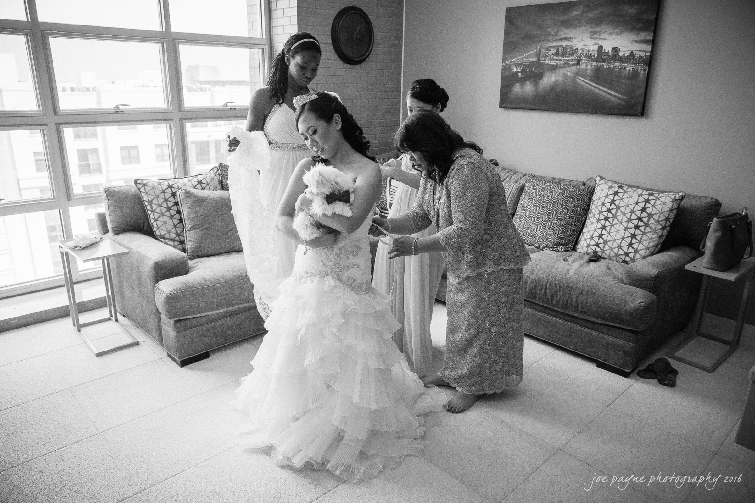A bride in a tiered gown holds a small dog while surrounded by three women helping her get ready. The room has a modern couch, large windows, and a cityscape picture on the wall.