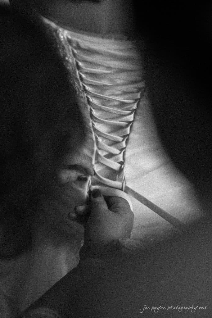 A black and white photo showing hands tightening the laces of a wedding dress. The image emphasizes the intricate pattern of the corset back and the care involved in the process.