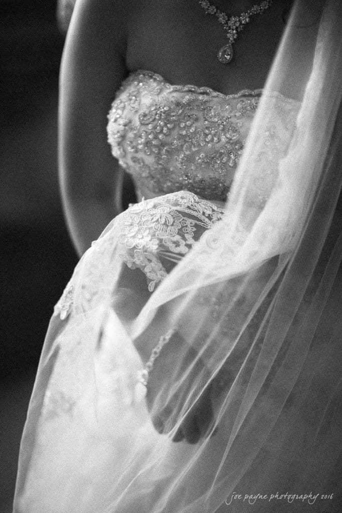 A black and white close-up of a bride in an ornate wedding dress with intricate beading and lace details, holding a delicate veil.