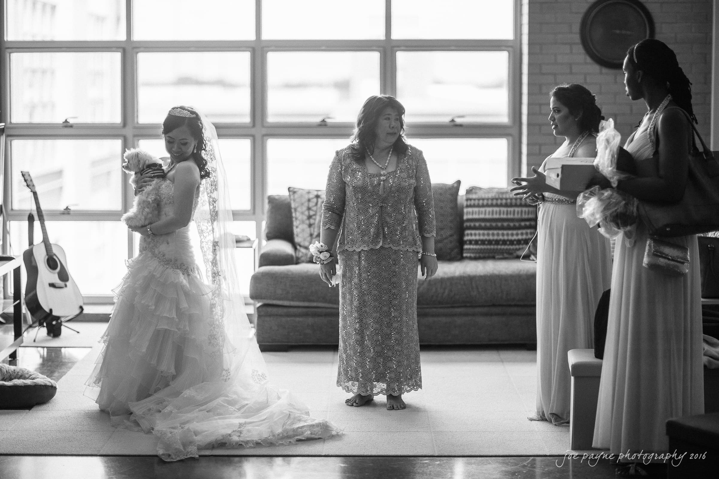 A bride in a wedding dress holds a small dog as she stands beside two elegantly dressed women in a room with a large window. A guitar rests against the wall.