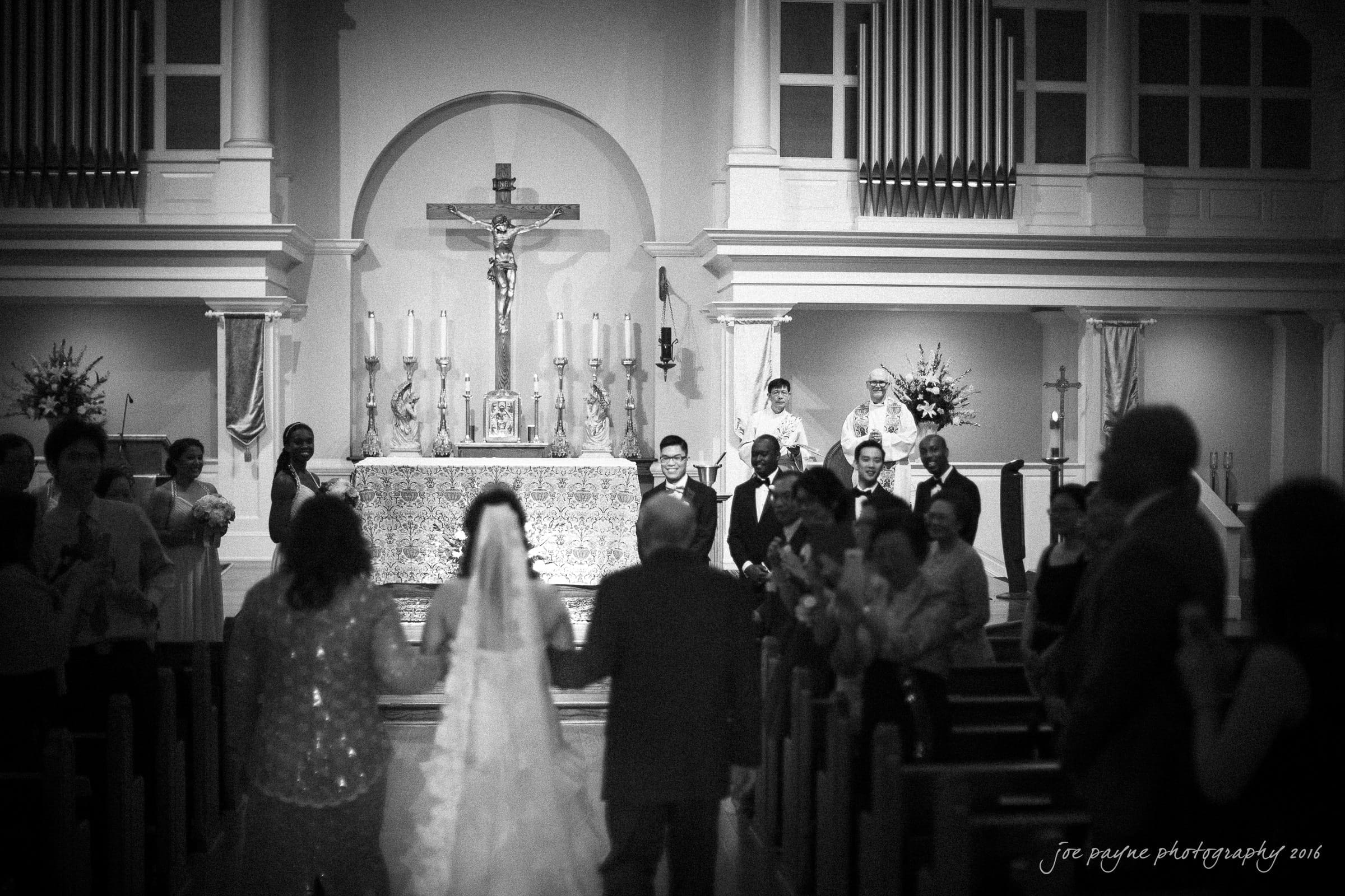 A bride and her parents walk down the aisle of a church toward the altar, where the groom and officiant await. Guests are seated on either side, some standing and clapping. The church is decorated with flowers and candles.