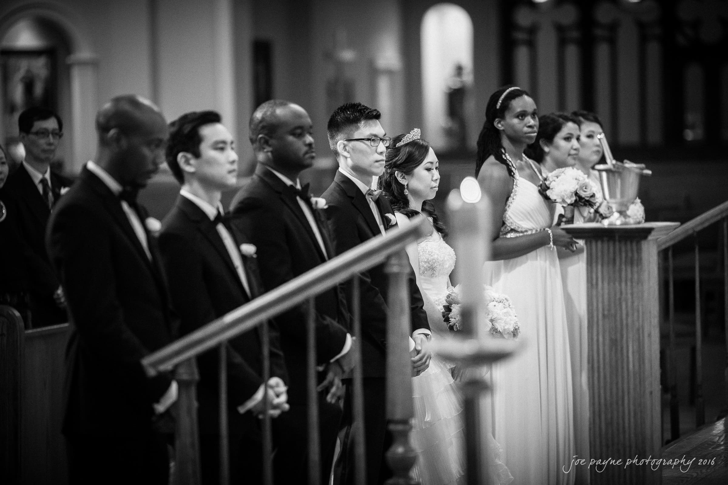 A black and white photo of a wedding ceremony in a church. The bride and groom stand at the altar with their bridal party. The wedding party is dressed in formal attire, and lit candles are visible in the foreground.