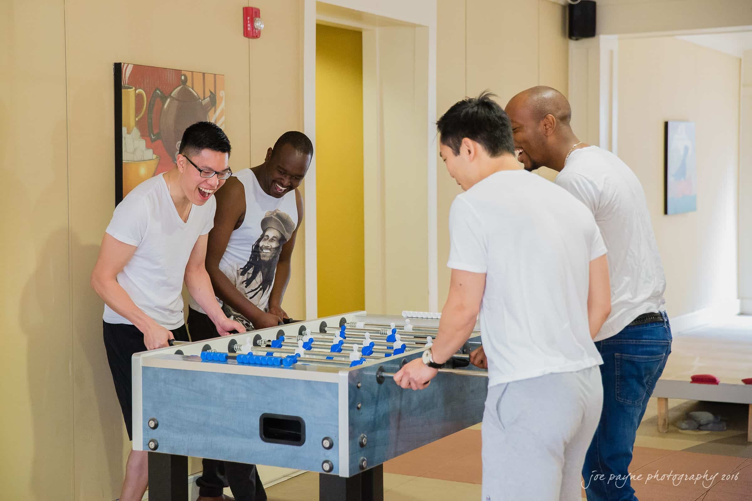 Four people are playing table soccer indoors, enjoying a lively game. Two are on each side of the foosball table, engaged and smiling. The room has a light-colored wall and artwork in the background.