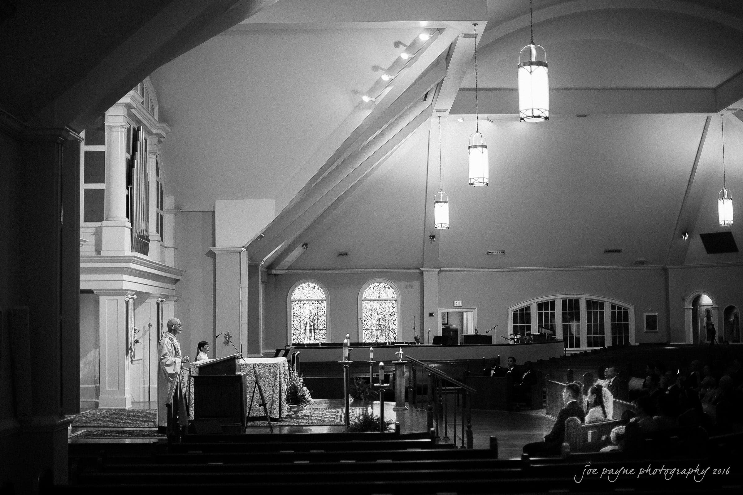 A black and white photo of a church interior with a priest standing at the altar. Rows of pews are partially filled with seated people. Stained glass windows and hanging lights illuminate the space.