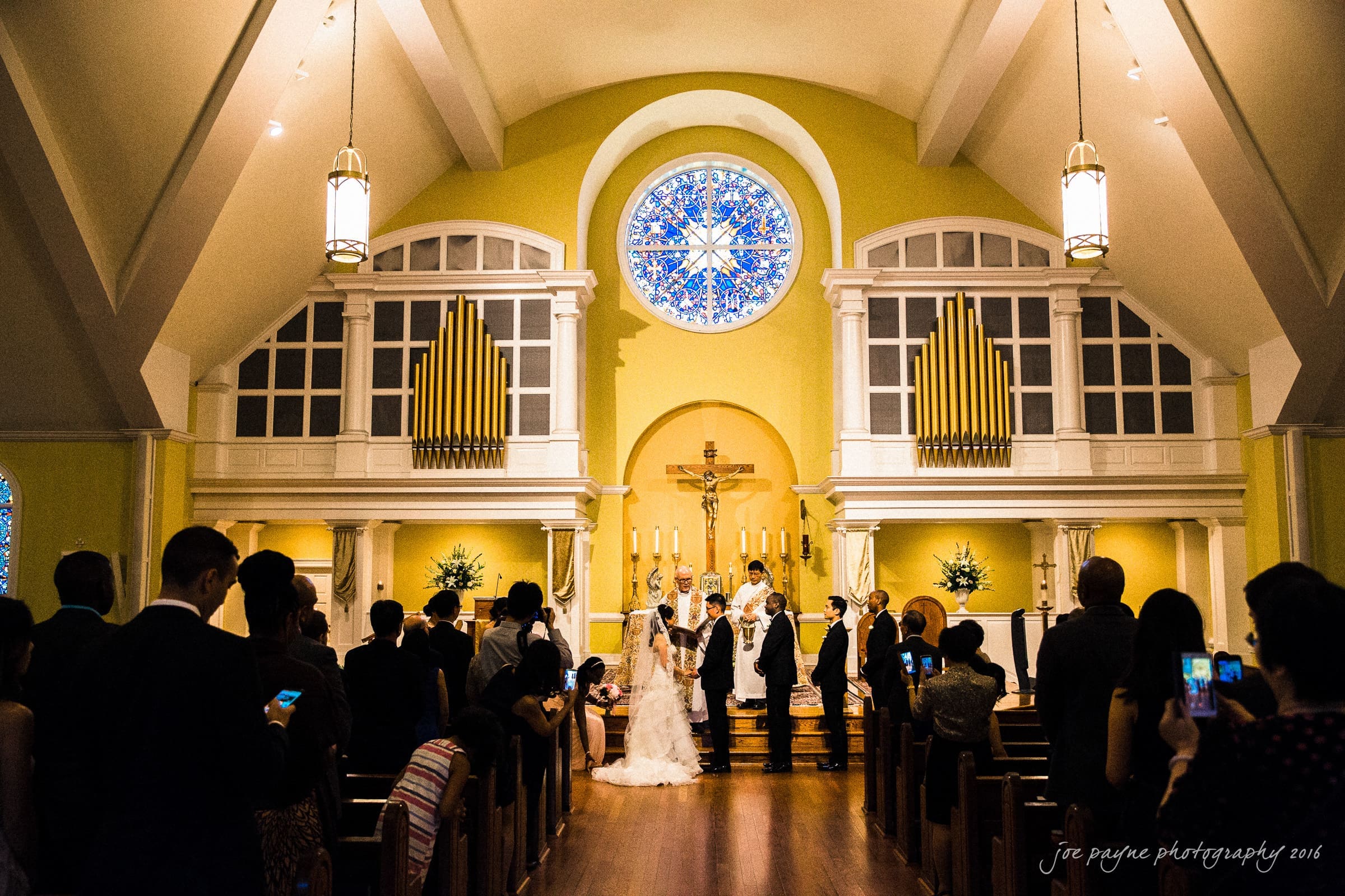 A wedding ceremony in a church with a high ceiling and stained glass window. The bride and groom stand at the altar, and guests are seated in pews, some taking photos. The interior features yellow walls and tall organ pipes.