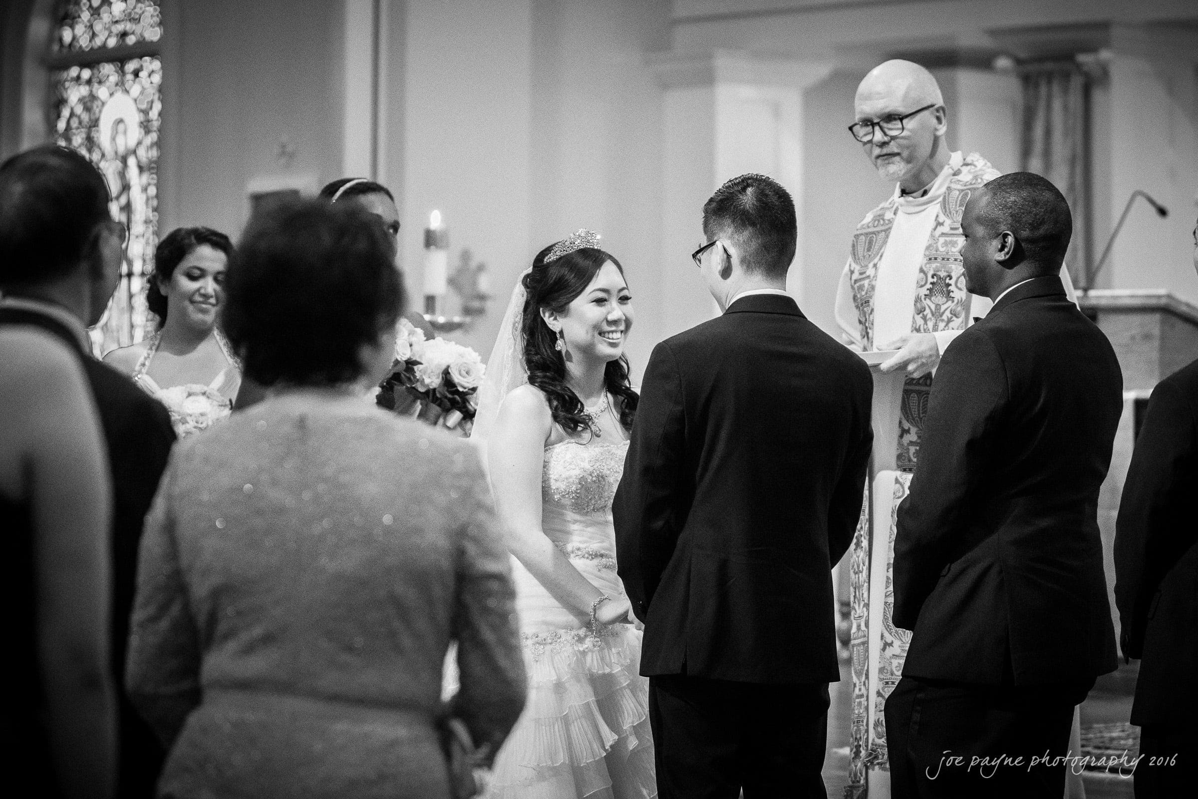 A bride and groom stand facing each other during a wedding ceremony inside a church. The bride is smiling, wearing a white gown and a tiara. A priest stands at the altar, while guests and bridal party members look on.
