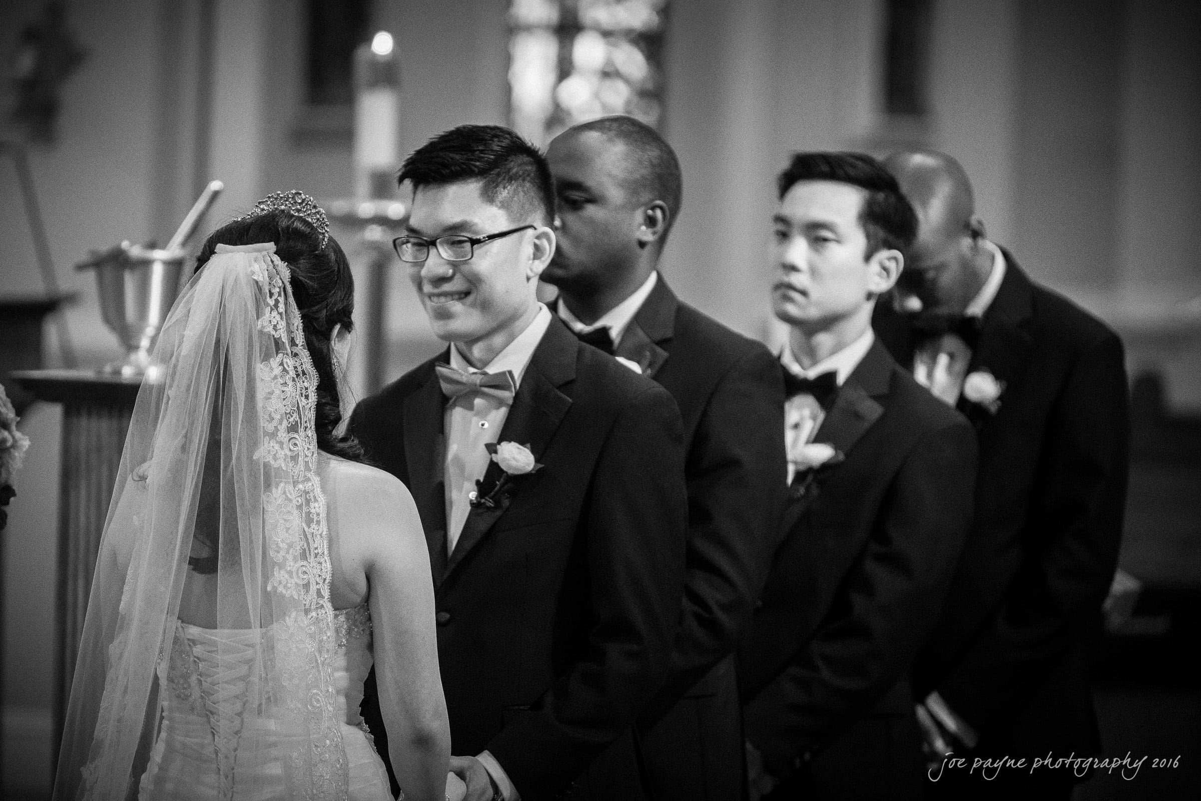 A bride and groom face each other and hold hands during their wedding ceremony. The groom and his groomsmen wear tuxedos with bow ties, and one holds his hand over his heart. The ceremony takes place in a church.