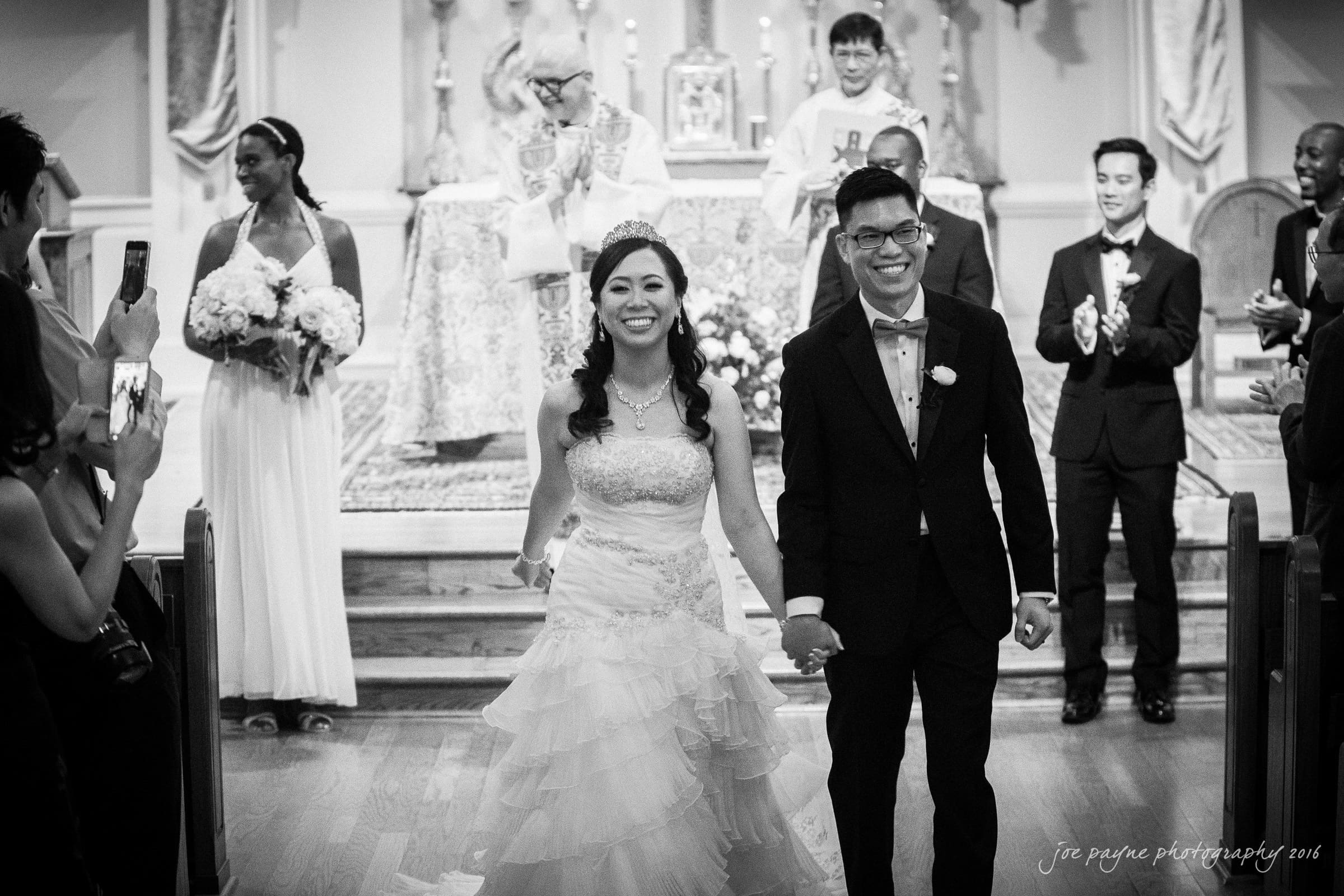 A bride and groom smile and hold hands while walking down the aisle. Bridesmaids and groomsmen stand in the background, with a priest at the altar. The wedding takes place inside a church.