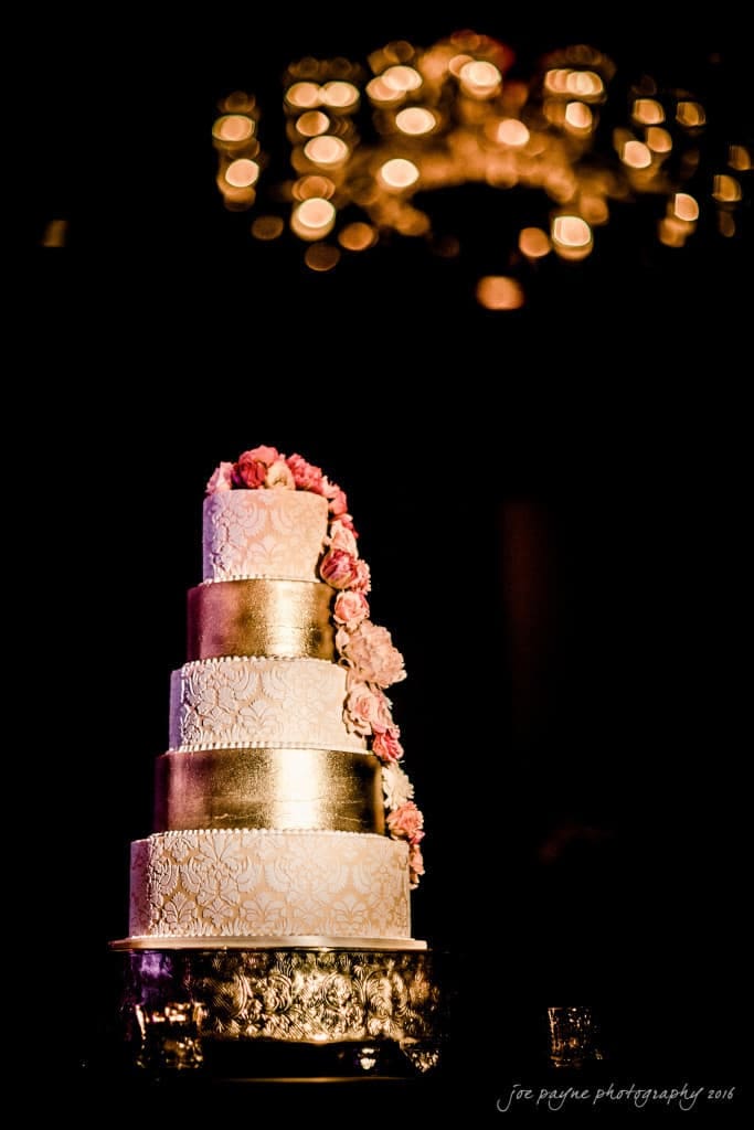 A four-tiered wedding cake decorated with intricate patterns and pink flowers cascading down one side. The tiers are silver and white. The cake sits on an ornate stand with a blurred, sparkling chandelier above.