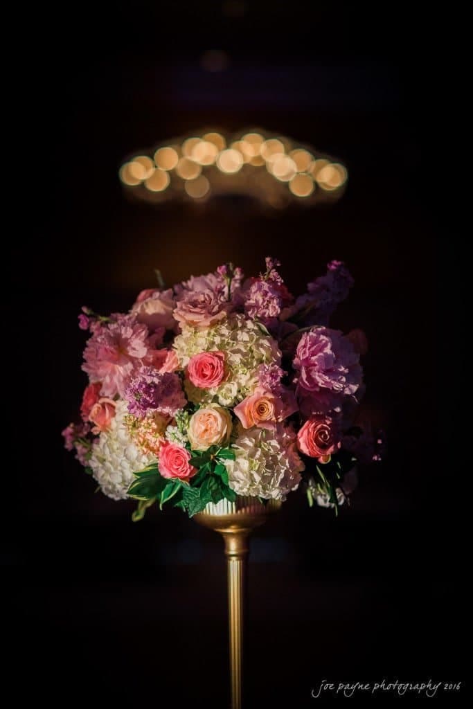 A bouquet of flowers, including pink roses, white hydrangeas, and purple blooms, sits on a tall stand against a dark background. Soft, blurred lights create a warm glow in the background.