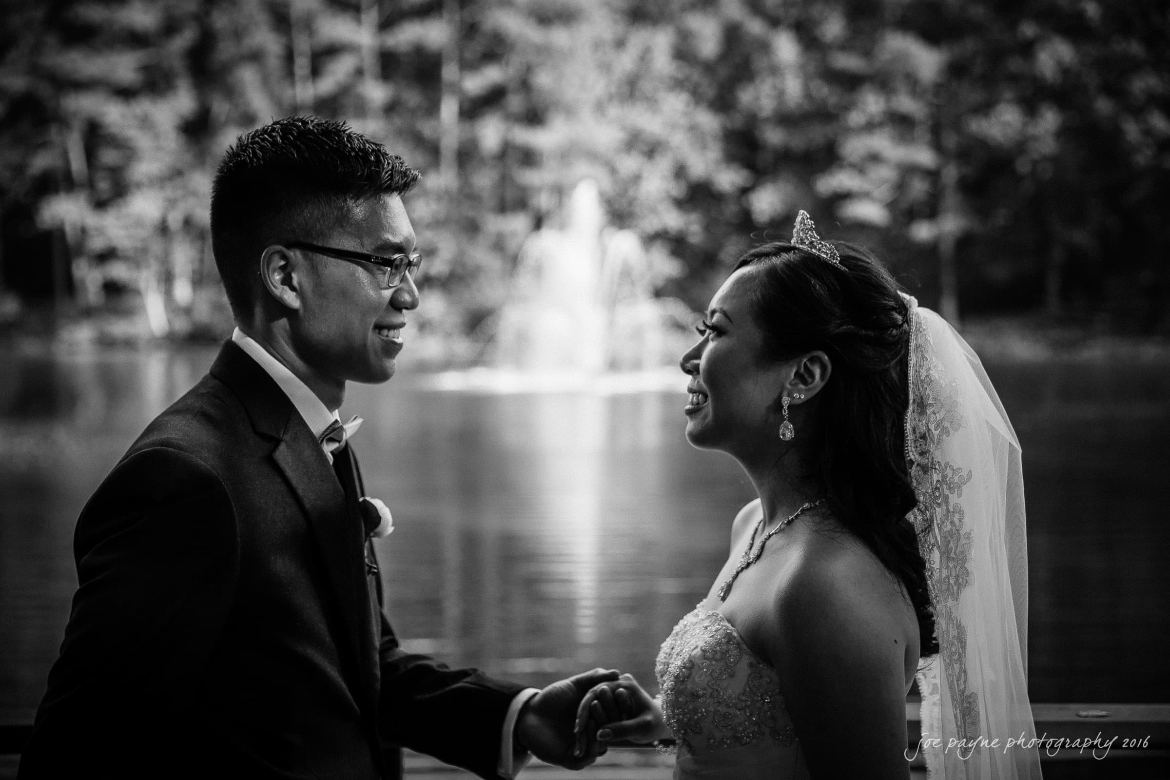 A bride and groom smiling at each other in front of a lake with a fountain. The bride is wearing a veil and tiara, while the groom is in a suit. The background is blurred, emphasizing the couples joyful interaction.