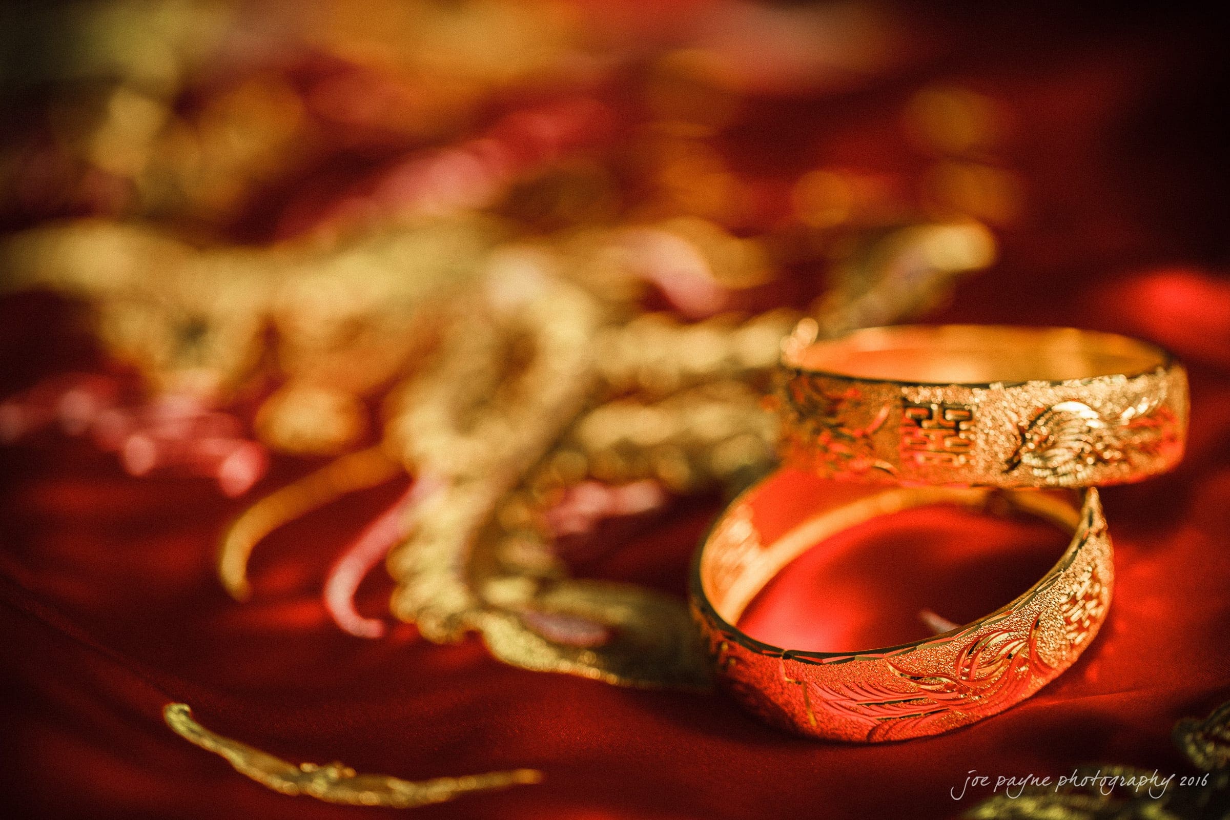 Two intricately designed gold bracelets rest on a vibrant red fabric adorned with golden embroidery. The background features soft, blurred patterns, emphasizing the elaborate details of the jewelry in the foreground.