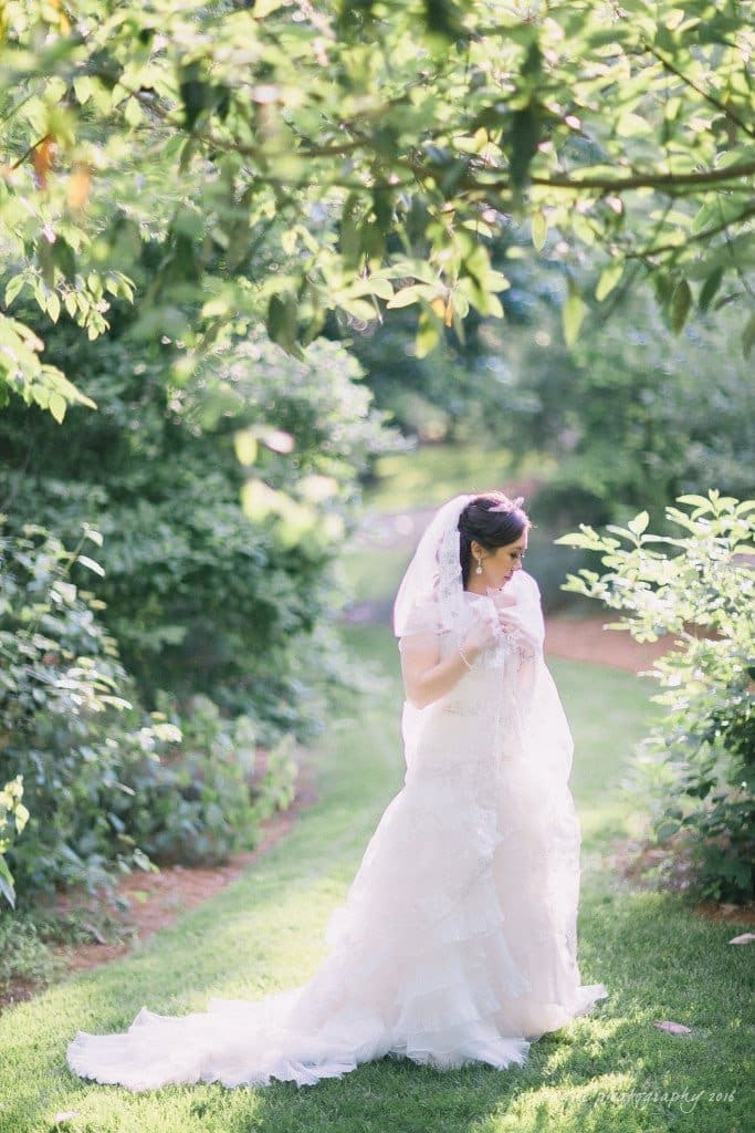 A bride in a white wedding dress and veil stands on a green grassy path surrounded by lush greenery. Sunlight filters through the leaves, creating a serene and romantic atmosphere.