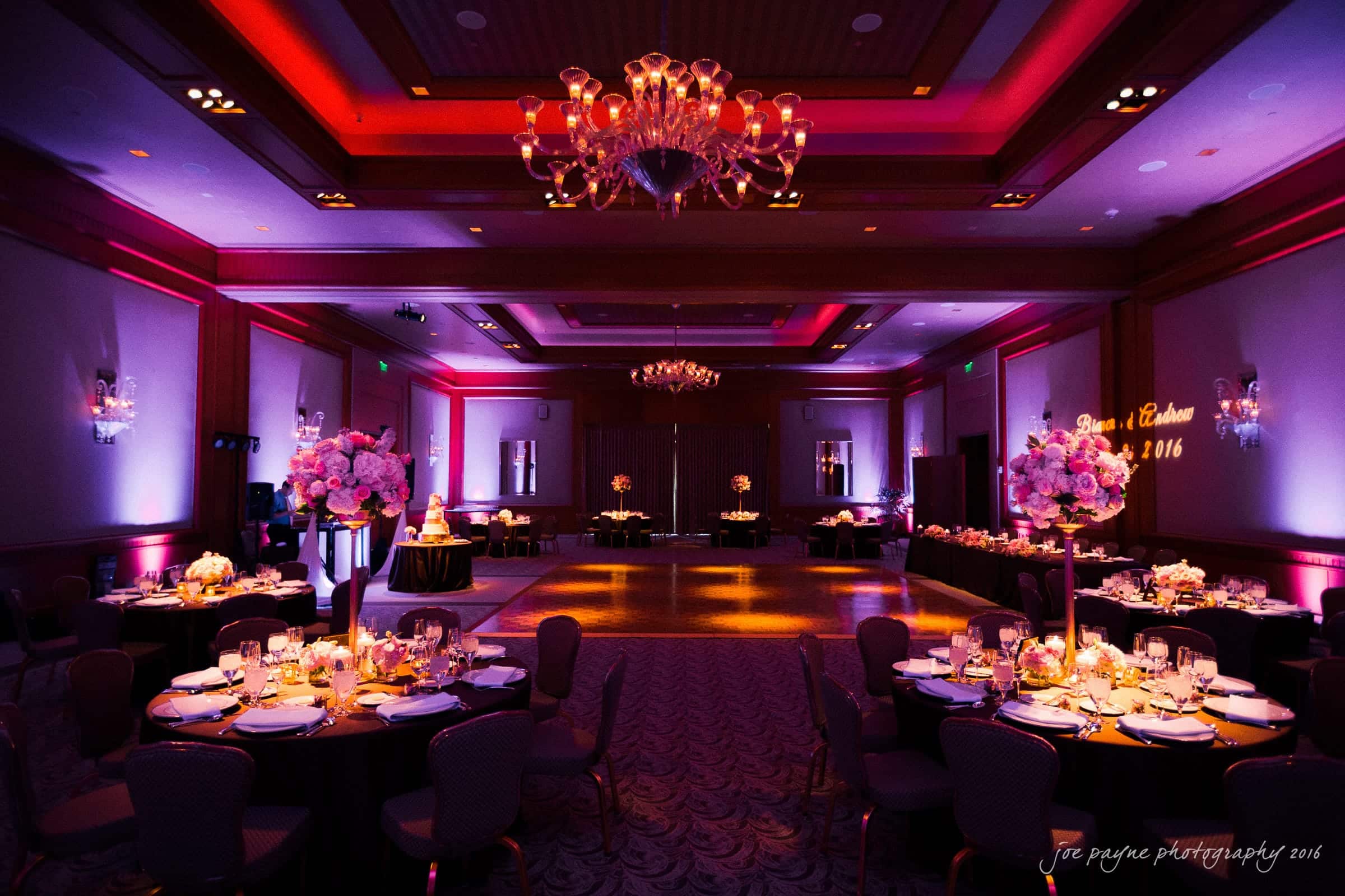 A dimly lit, elegantly decorated ballroom with tables set for a formal event. Each table features floral centerpieces and candles. The room has chandeliers and pink and purple lighting that enhances the ambiance. A dance floor is visible in the center.