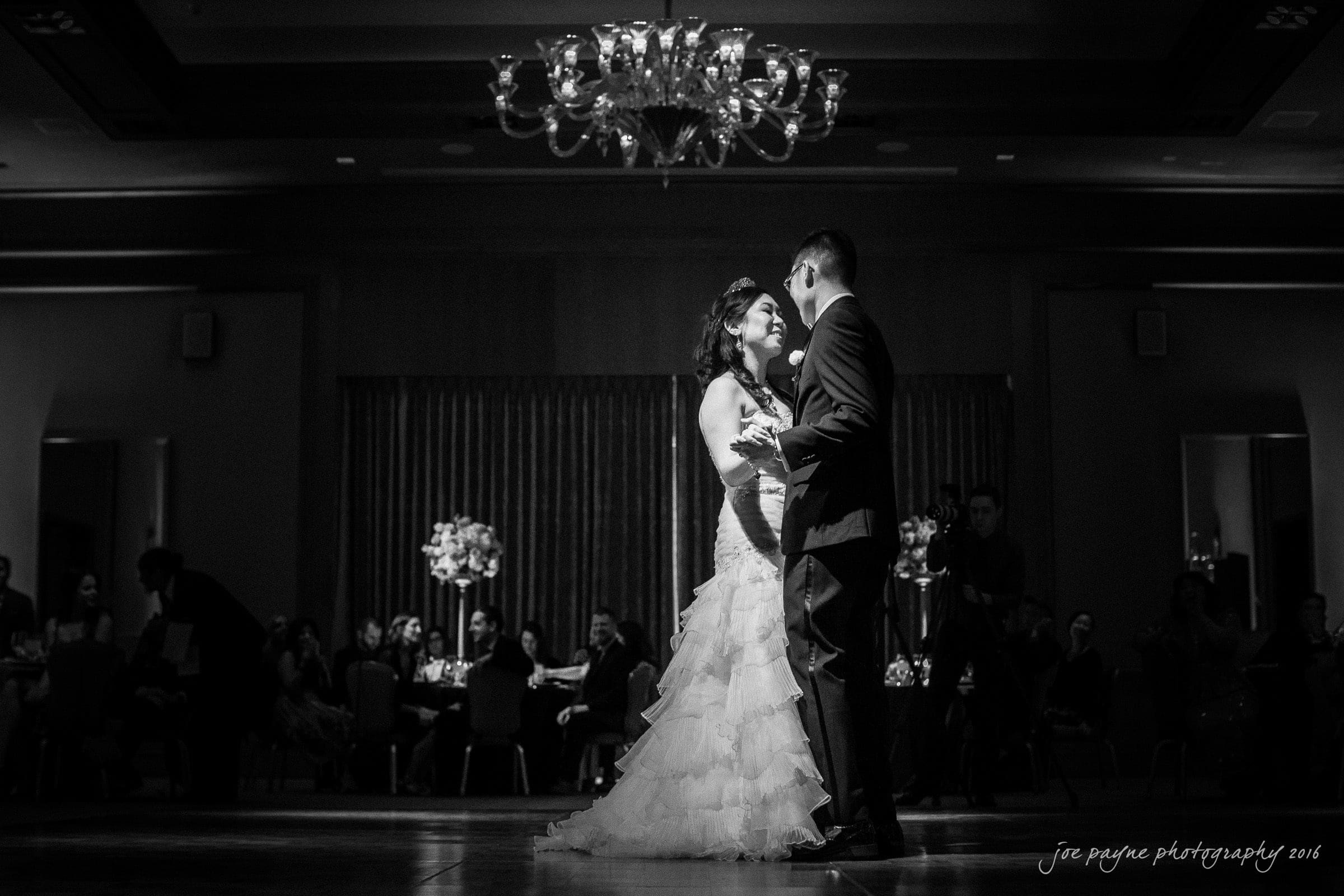 A bride and groom share their first dance in an elegantly lit ballroom. The room is dimly lit except for a spotlight on the couple. Guests watch from their tables, and a chandelier hangs above.