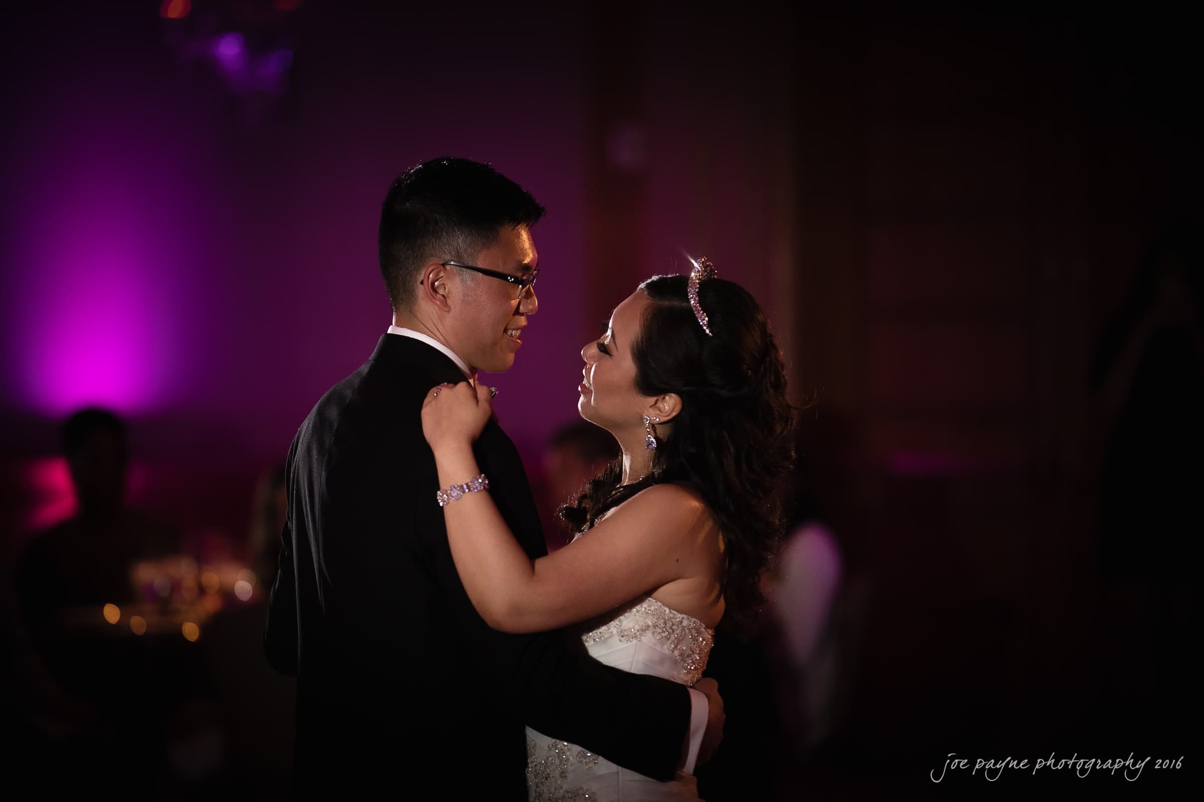A bride and groom share a first dance at their wedding. The groom wears a black suit, and the bride wears a white gown and tiara. The background is dimly lit with purple lighting, creating a romantic atmosphere.