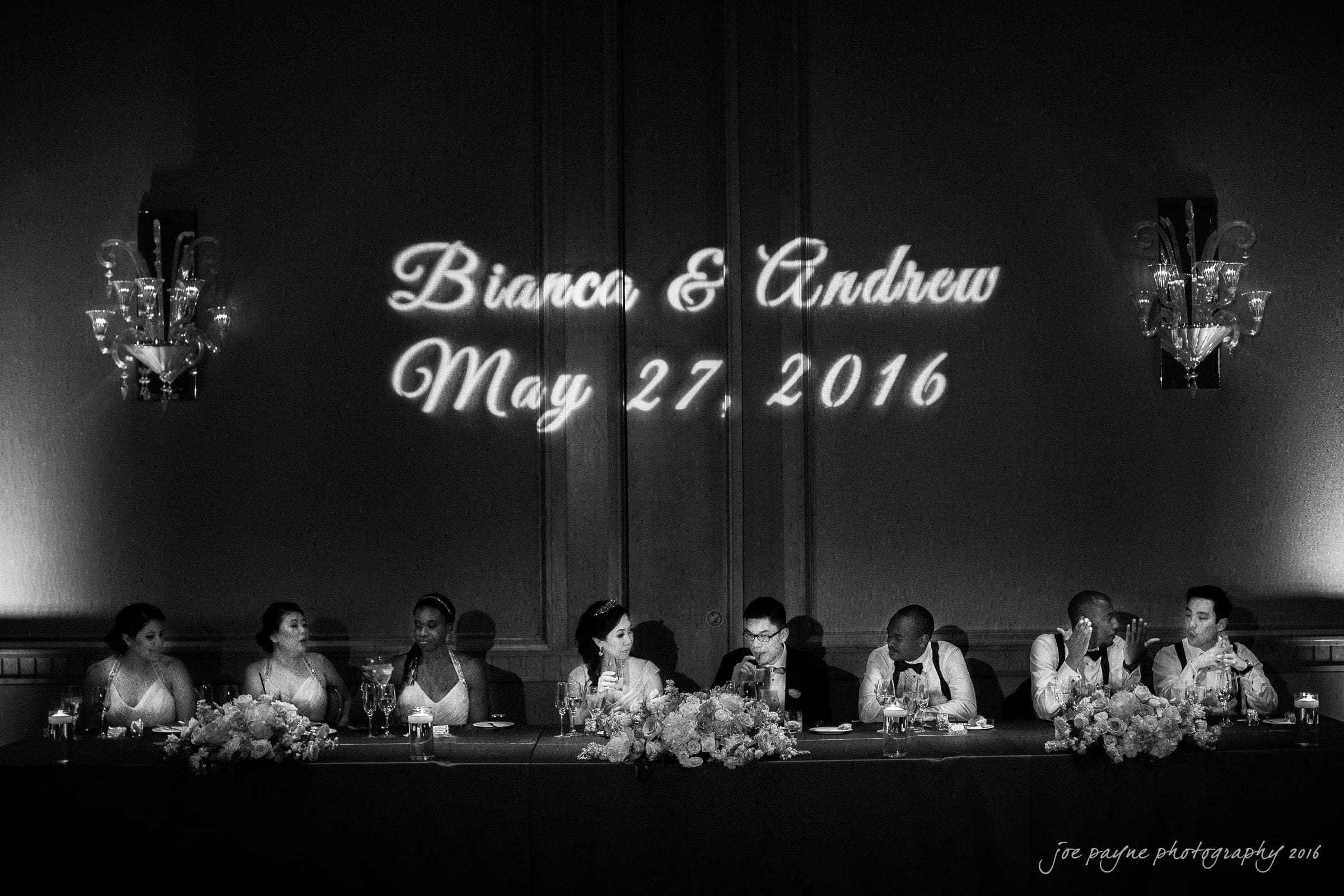 A black and white photo of a wedding reception. Nine people are seated at a long table decorated with flowers and candles. Above them, Bianca & Andrew, May 27, 2016 is illuminated on the wall. Elegant chandeliers are visible on both sides.