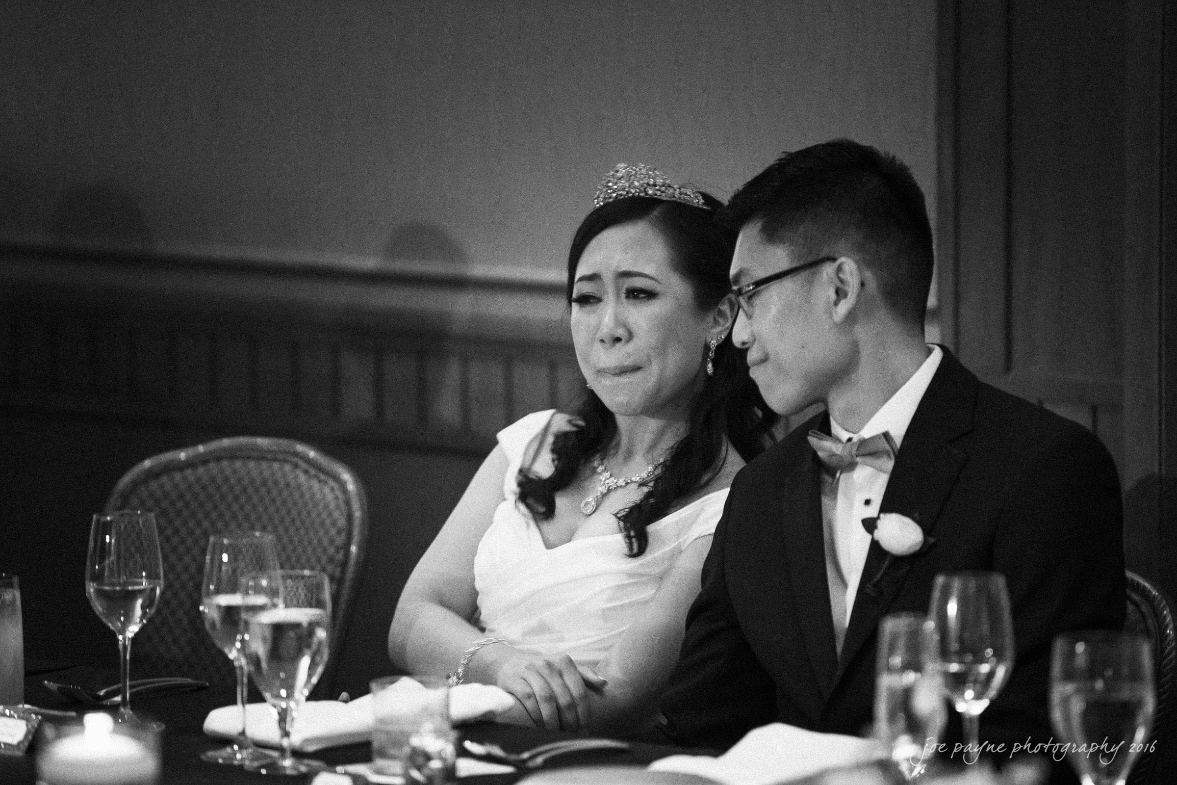 A bride and groom sit closely at a table. The bride looks emotional, wearing a tiara and white dress, while the groom in a suit leans in supportively. Glasses and candles are on the table, creating an intimate atmosphere. Black and white photograph.