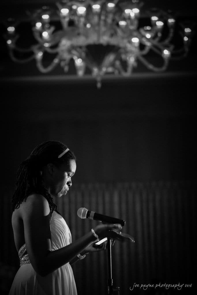 A woman in a white dress speaks into a microphone under a large chandelier. The setting is elegant and dimly lit, creating a dramatic atmosphere.