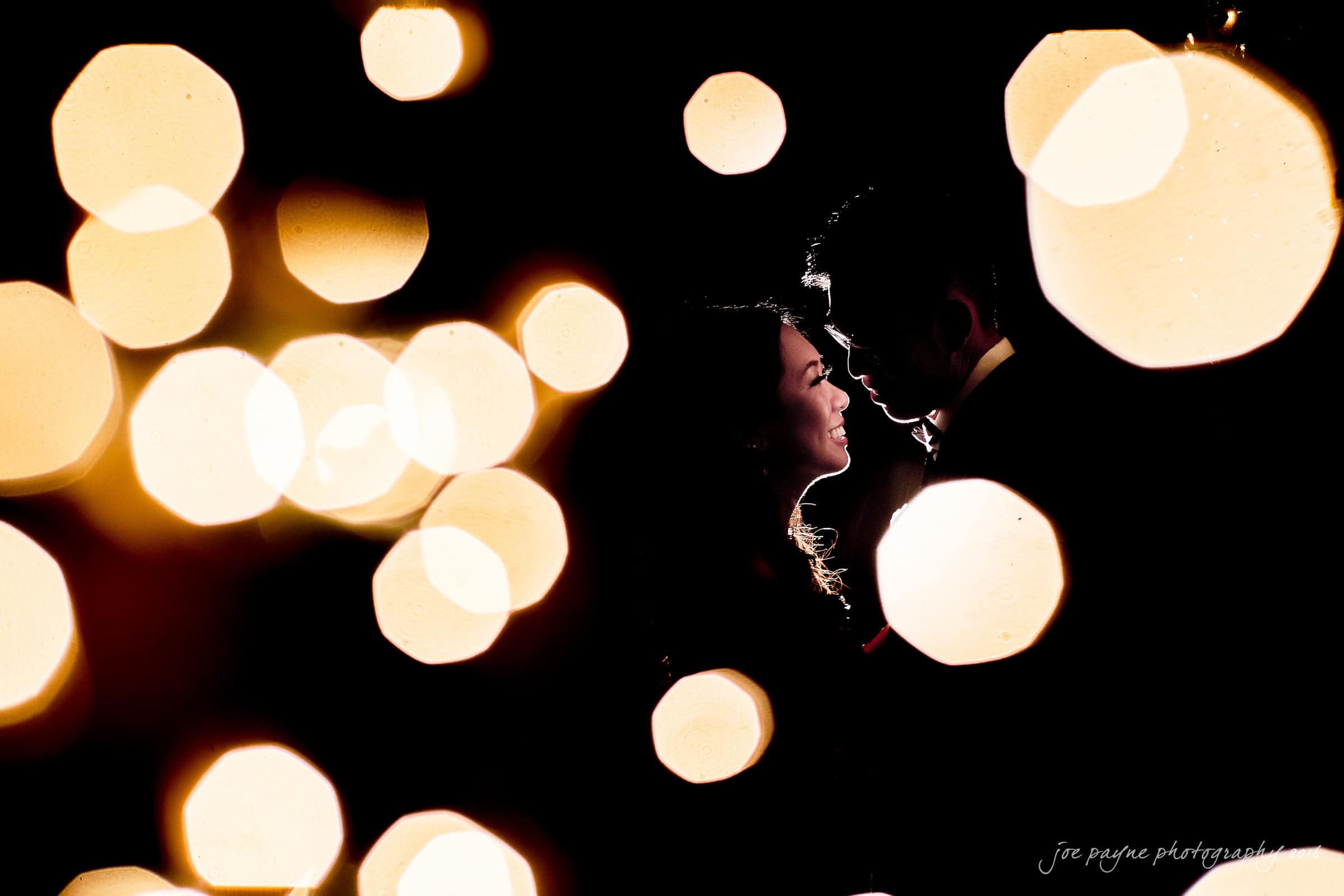 A silhouetted couple faces each other, smiling tenderly, surrounded by large, blurred golden lights against a dark background, creating a romantic and dreamy atmosphere.