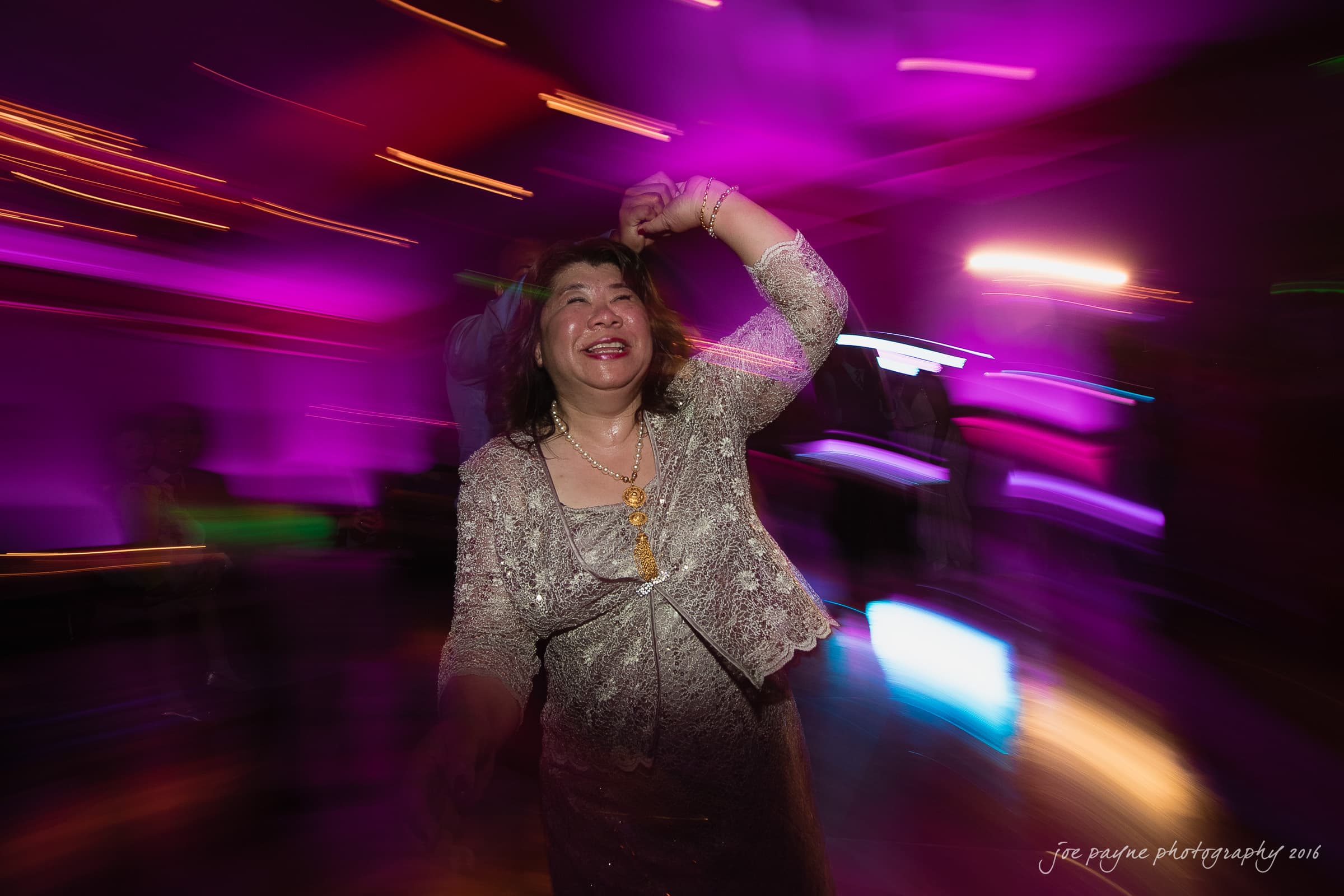 A woman joyfully dances in a vibrant, colorful setting with blurred lights and motion effects. She is smiling widely, wearing a sparkly outfit, and holding her arm up in celebration.