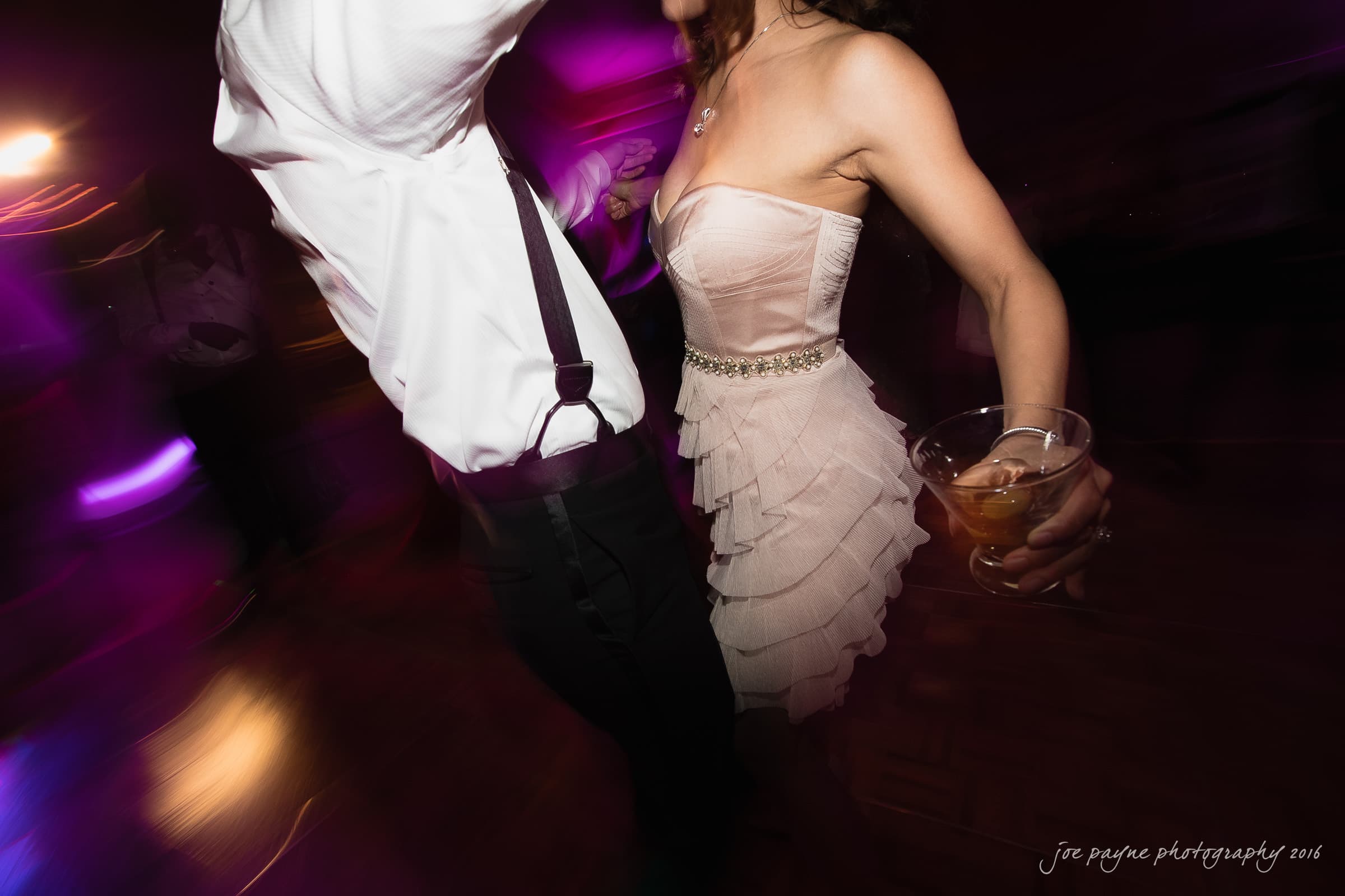 A couple is dancing energetically in a dimly lit room. The person on the right is wearing a strapless, ruffled dress and holding a drink. The mood is lively, and the colorful lights add a dynamic, swirling effect to the scene.