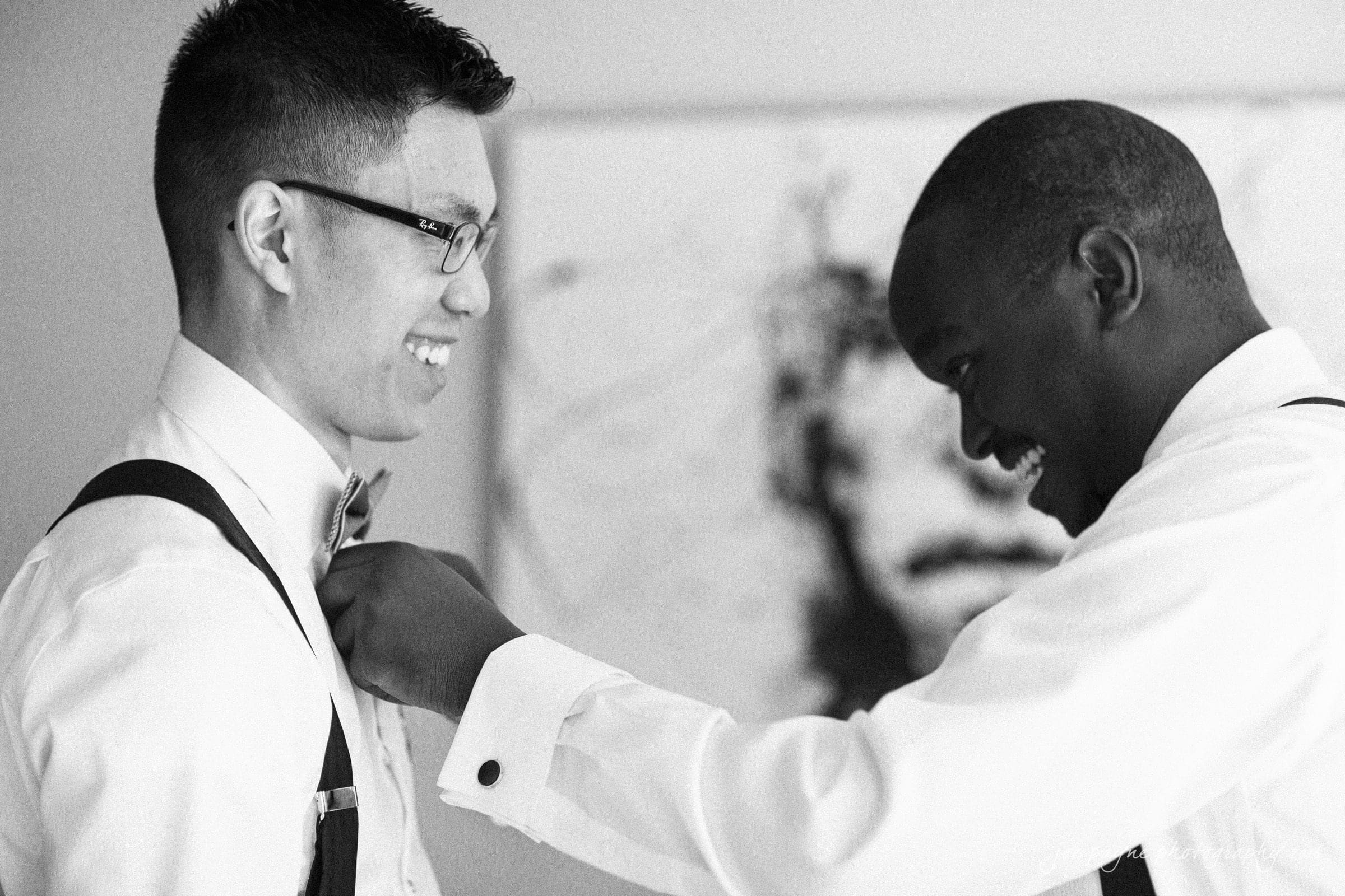 A black-and-white photo of two men in formal attire. One man is helping the other adjust his bow tie. They are smiling and look happy. A blurred painting is visible in the background.