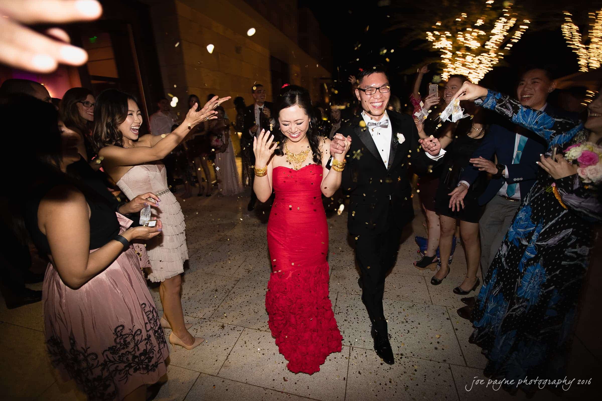 A couple in formal attire, the woman in a red dress and the man in a black suit, walk jubilantly through a crowd. Guests are joyfully tossing confetti at them, celebrating in a warmly lit evening setting.