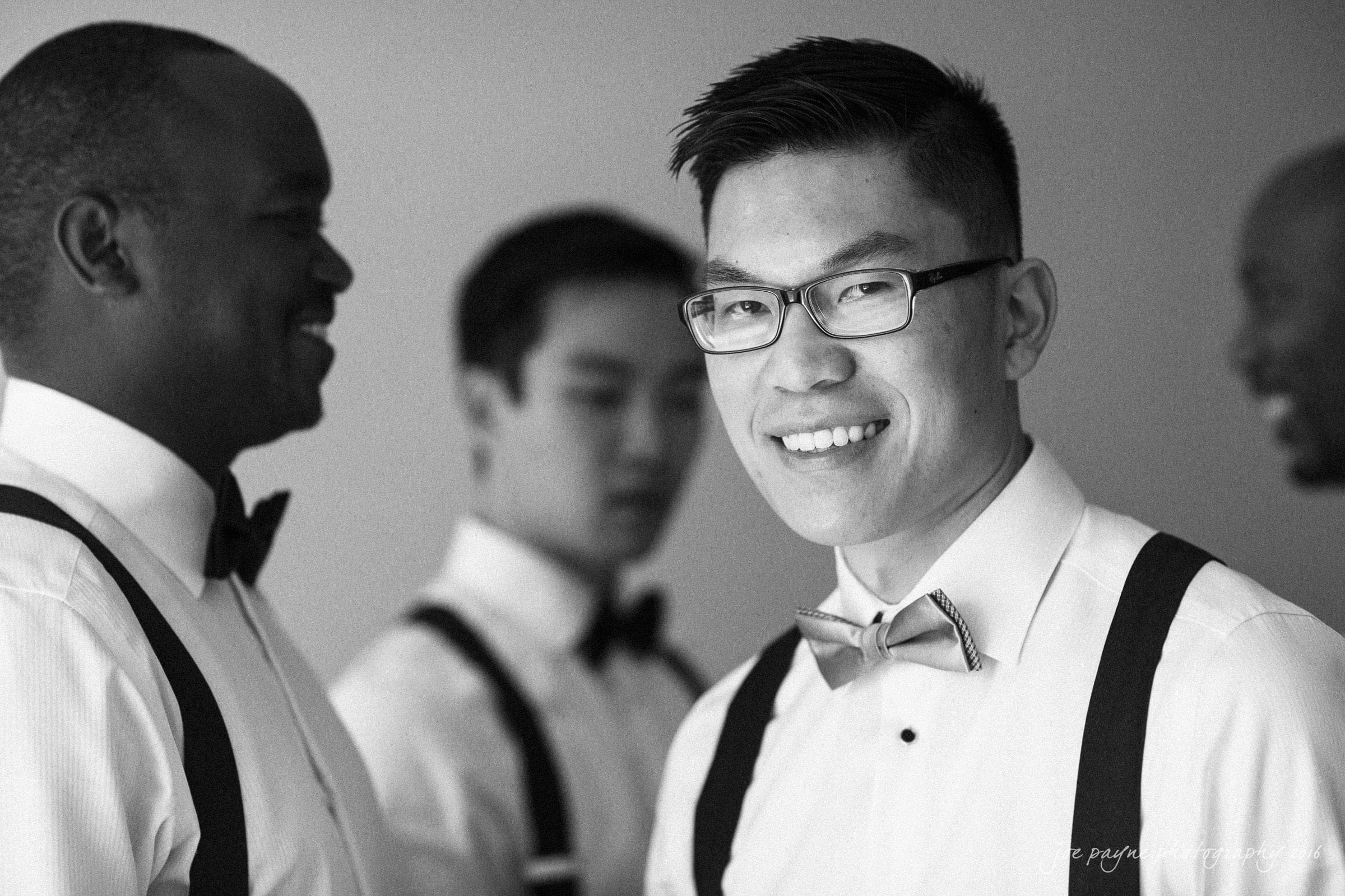 Four men in formal attire and bow ties are gathered together. One man in the foreground is smiling at the camera, while the others are engaged in conversation in the background. The image is black and white.