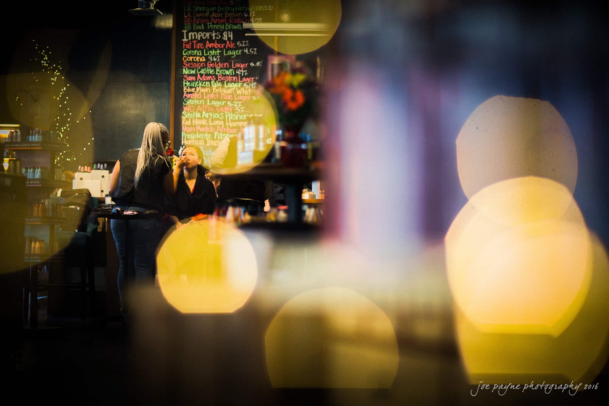 A cozy coffee shop with two people chatting near a wall menu. Soft, warm lighting and decorative bokeh effects give the scene a relaxed ambiance. A vase with flowers is visible on a counter.