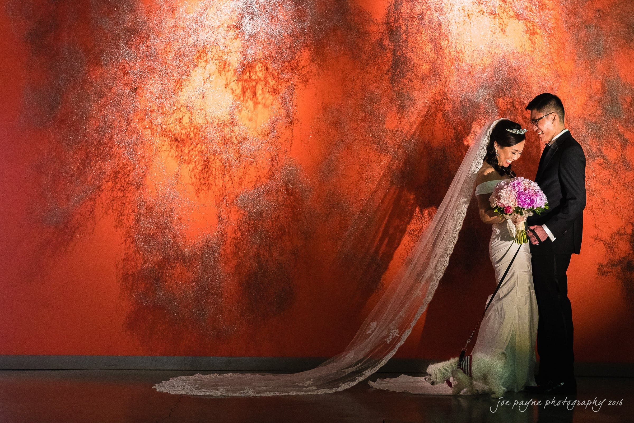 A bride and groom stand close together in front of an orange textured wall. The bride, in a long white dress and veil, holds a bouquet of purple flowers. The groom is in a suit, smiling at her. Soft lighting casts a warm glow on them.