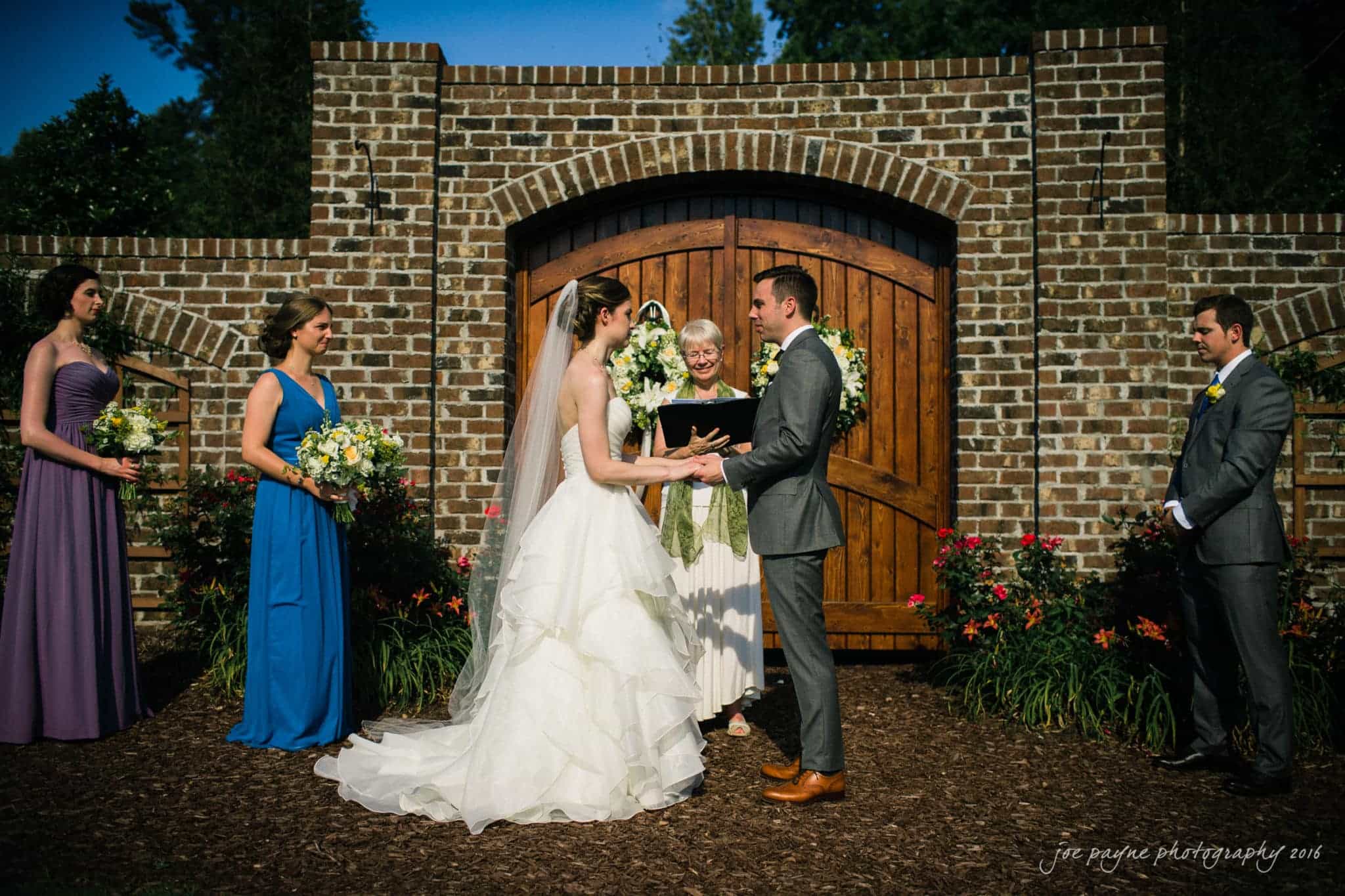 the oaks at salem wedding photography - kathryn & justin A bride and groom exchange vows in an outdoor wedding ceremony. They stand before a brick arch with wooden doors. Three bridesmaids and an officiant are present, each holding flowers. The scene is sunny and surrounded by greenery.