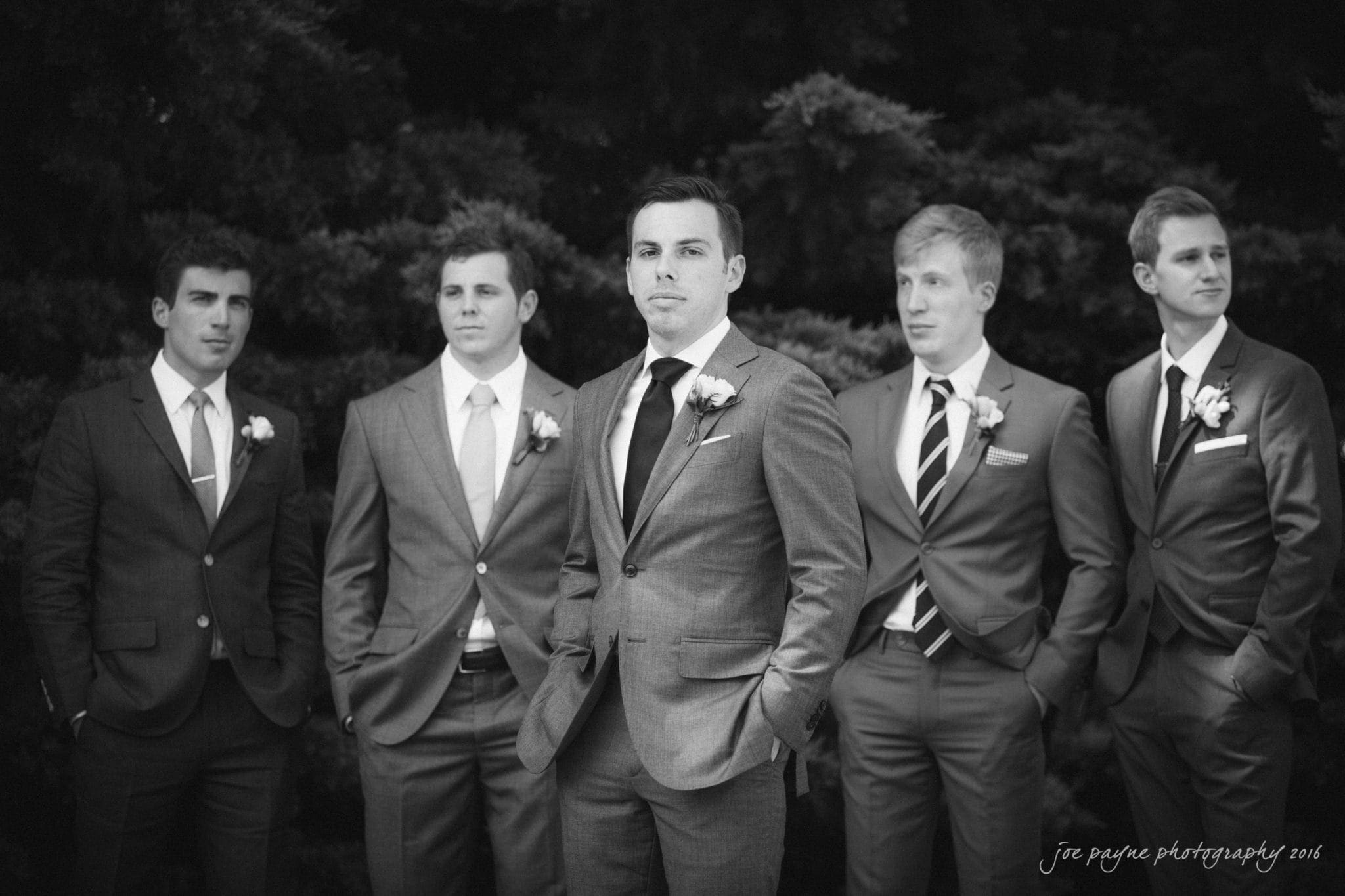 the oaks at salem wedding photography - kathryn & justin A black and white photo of five men standing outdoors, wearing suits with boutonnieres. They are posed casually, with the central figure in focus, hands in pockets. The backdrop is a dark, leafy setting.