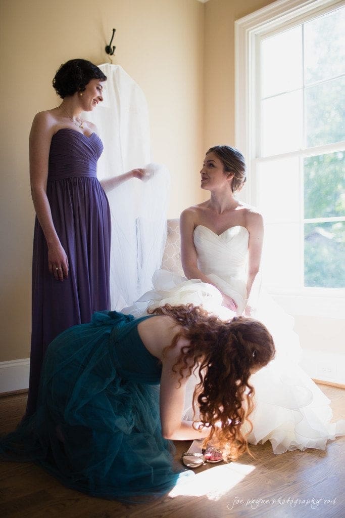 the oaks at salem wedding photography - kathryn & justin A bride in a white gown sits by a large window, looking at a woman in a purple dress who holds her veil. Another woman in a teal dress adjusts the brides dress. Natural light fills the room.