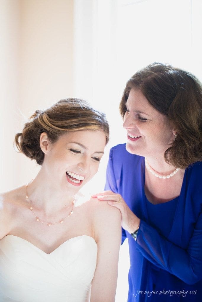 the oaks at salem wedding photography - kathryn & justin A bride in a white dress and a woman in a blue outfit share a joyful moment. The woman gently places her hand on the brides shoulder while they both smile warmly. Soft natural light illuminates them from a nearby window.
