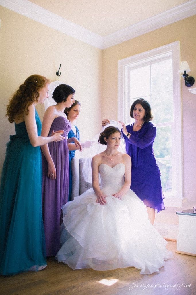 the oaks at salem wedding photography - kathryn & justin A bride in a white dress sits while smiling women in colorful dresses adjust her veil. The room is bright with natural light streaming in from a large window.
