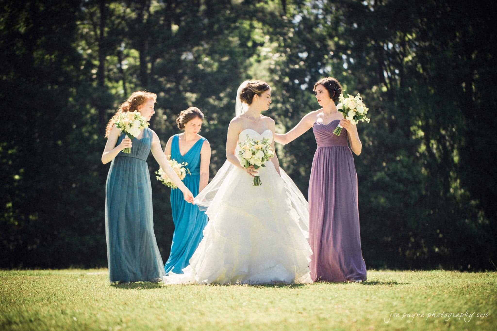 the oaks at salem wedding photography - kathryn & justin Four women in formal dresses stand on grass with trees in the background. The bride, in a white gown, holds a bouquet as sunlight filters through the trees. Bridesmaids in purple and teal dresses surround her, each holding similar bouquets.