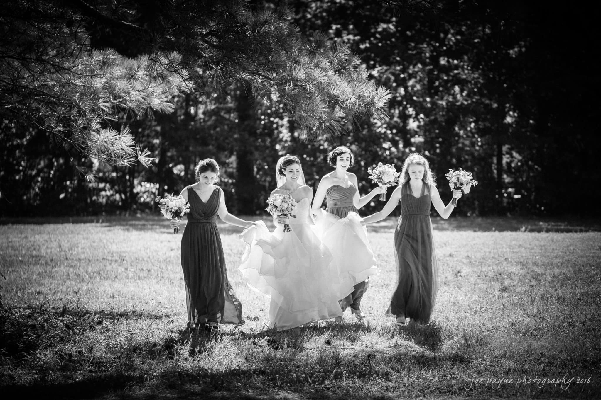 the oaks at salem wedding photography - kathryn & justin A black and white photo of four women walking in a grassy area holding bouquets. Theyre wearing elegant dresses, with the central figure in a long gown. Dappled sunlight filters through the trees, casting shadows on the ground.